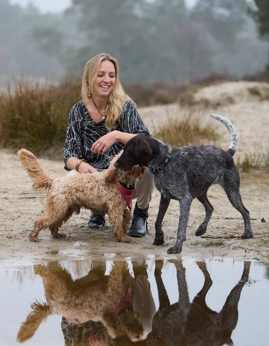 Vrouw knielt op het strand en speelt met twee honden, een met een krullige vacht en een met een speckled vacht, terwijl ze naar hun reflectie in het water kijkt.