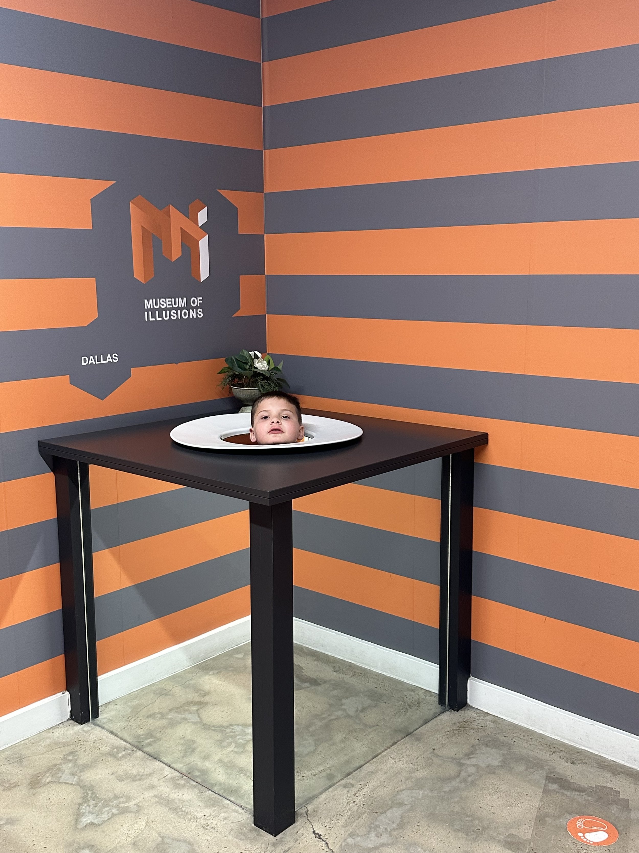 Museum of Illusions exhibit with a child's head on a table, creating an optical illusion, against a gray and orange striped background.
