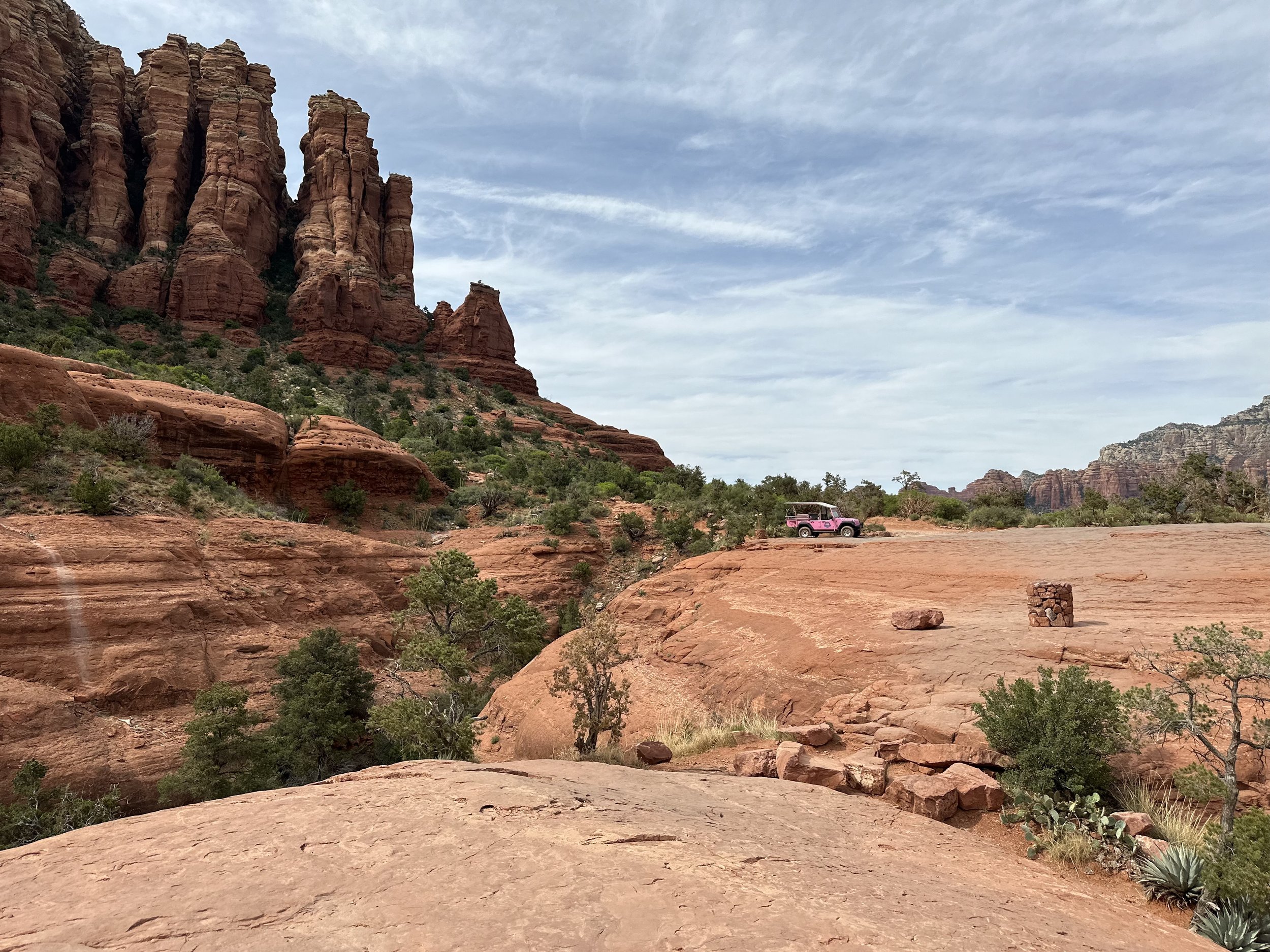 Red rock formations and landscape with a pink Jeep on a plateau in Sedona, Arizona, under a cloudy sky.