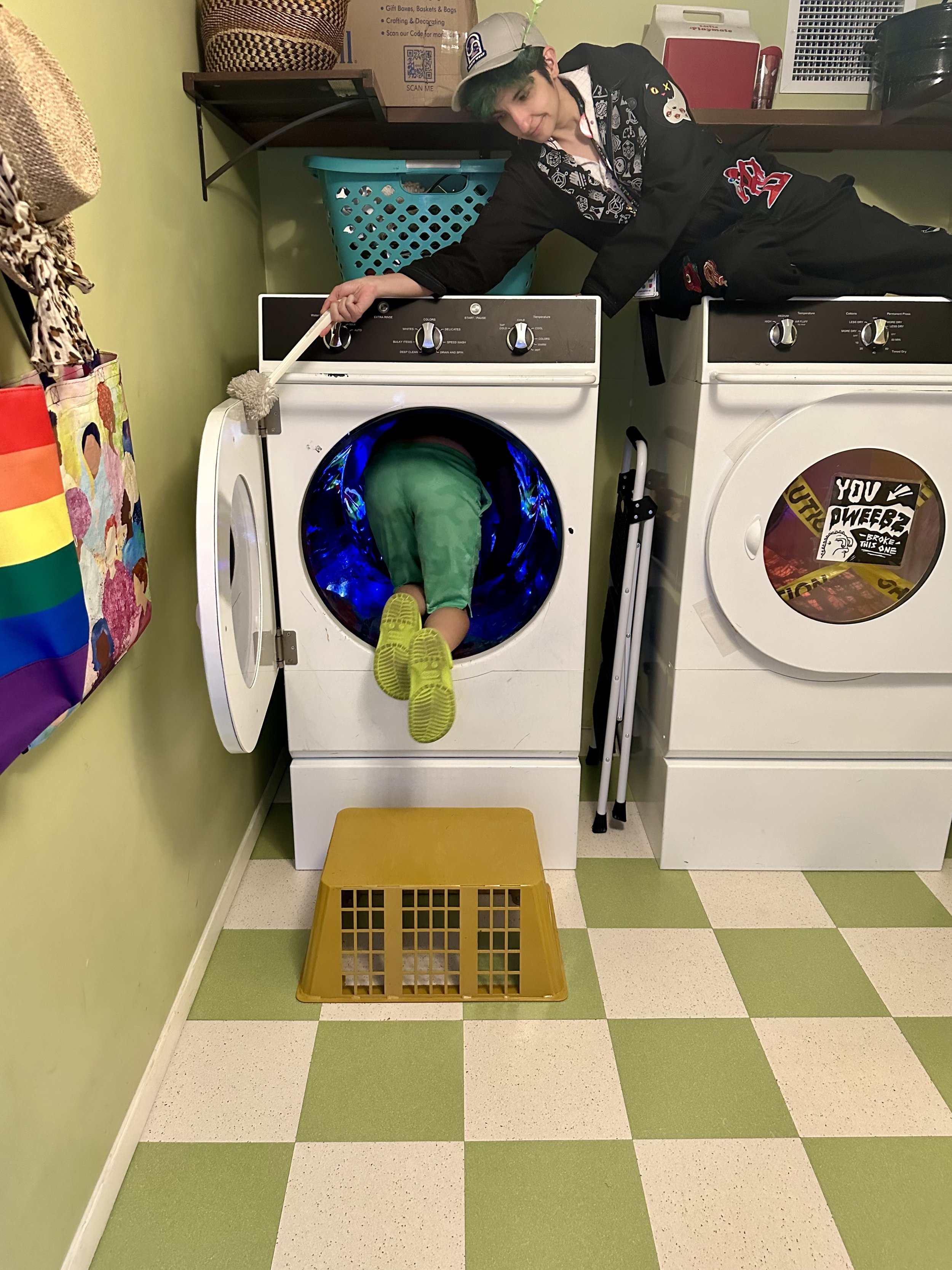 Two people in a laundry room. One person is inside a front-loading washing machine, legs sticking out, wearing green pants and shoes. Another person, wearing a hat and dark clothing with patches, is leaning over the machine, holding a brush. A laundr