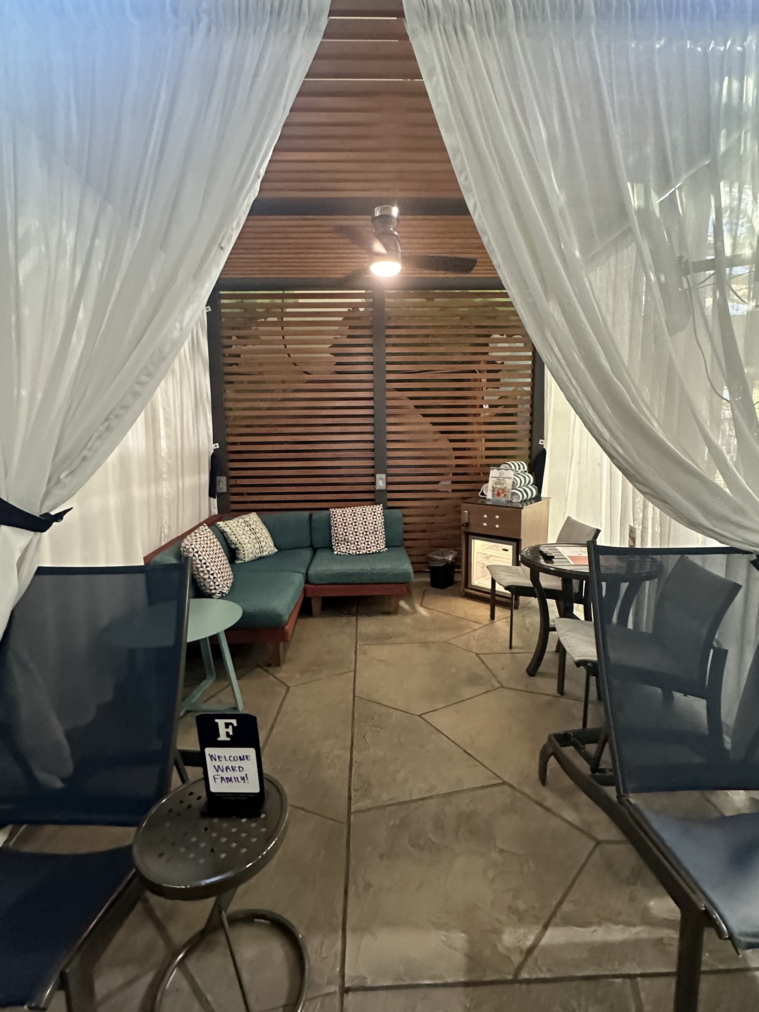 Outdoor cabana with white curtains and patio furniture, including a sofa, chairs, and tables, alongside a small sign reading 'Welcome Ward Family.'"