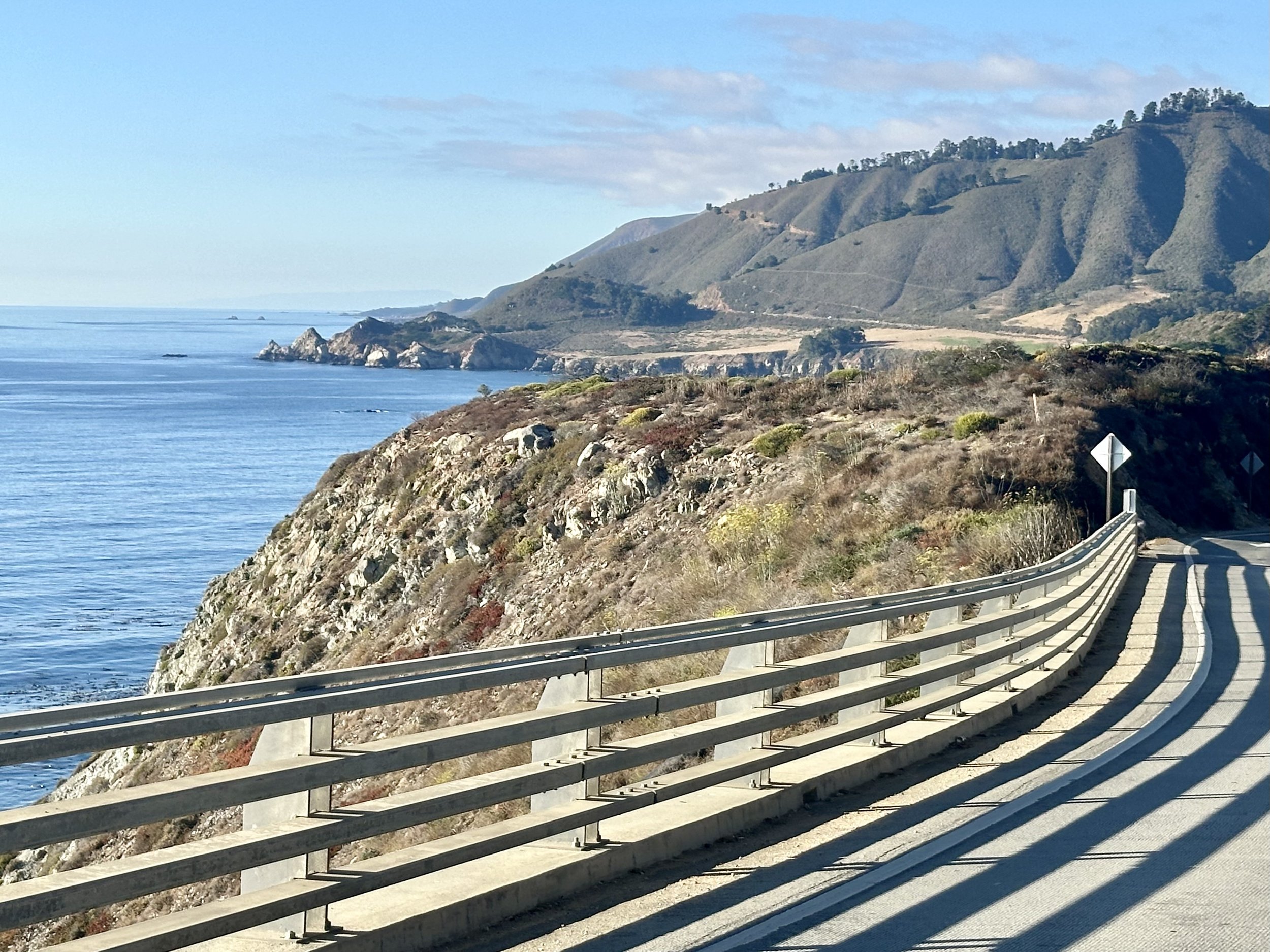 Coastal highway along a cliff overlooking the ocean in Big Sur, California, with mountains in the background.