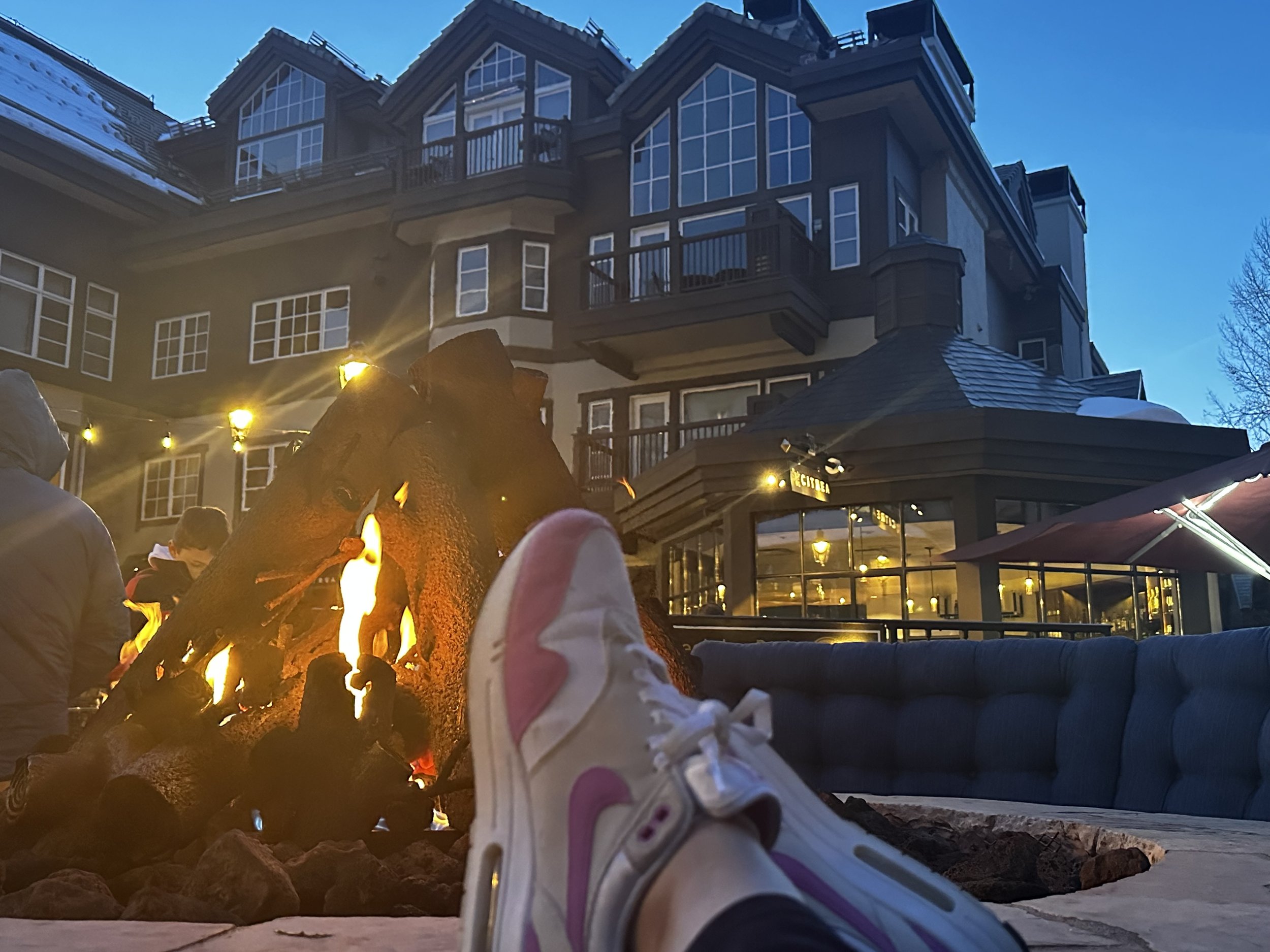 Outdoor fireplace with burning log at evening, person wearing white and pink sneakers with feet up, large multi-story building in background, warm light creating cozy atmosphere.