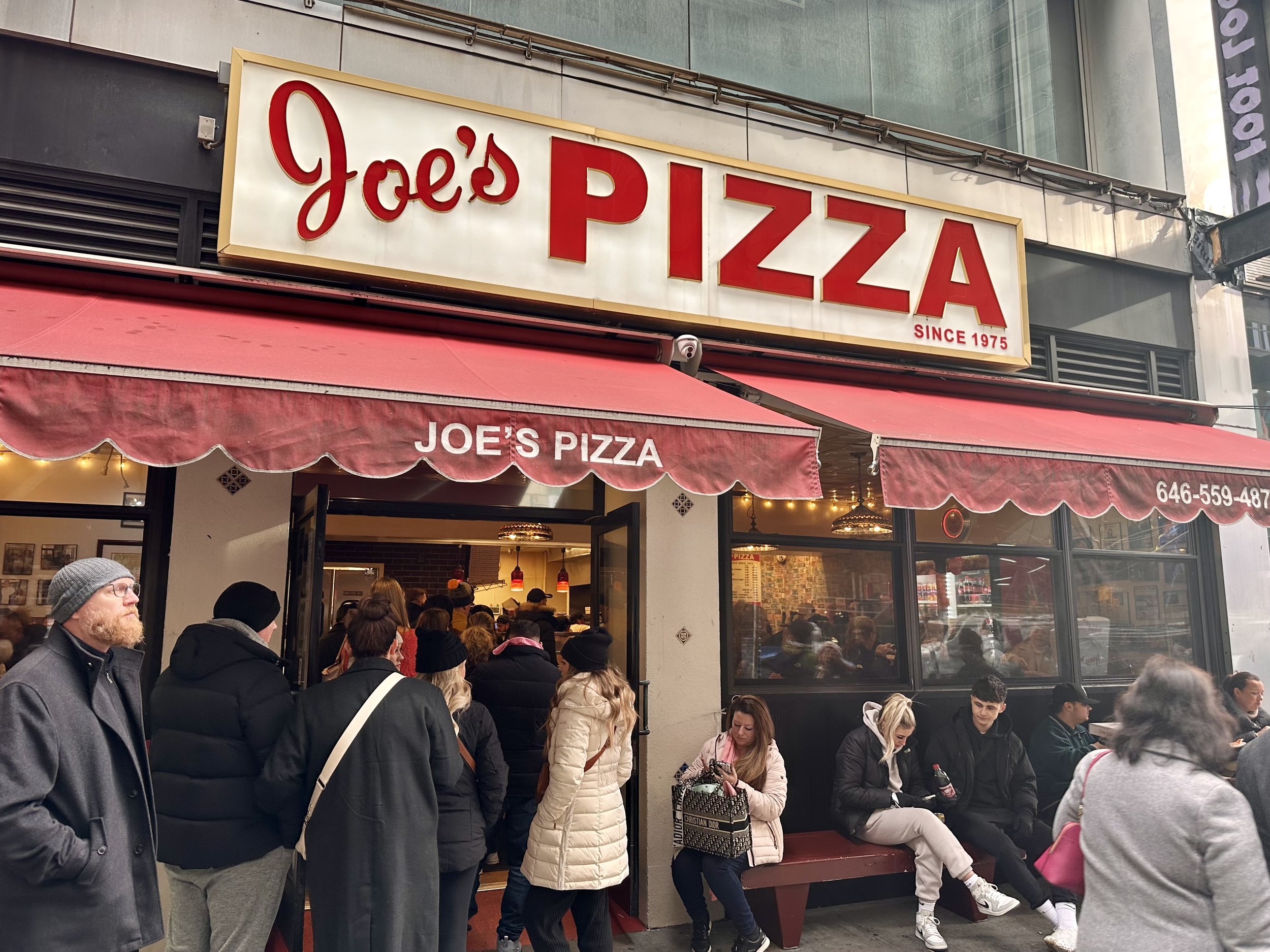 Exterior of Joe's Pizza with red awning, featuring people lined up outside the entrance and seated on a bench.