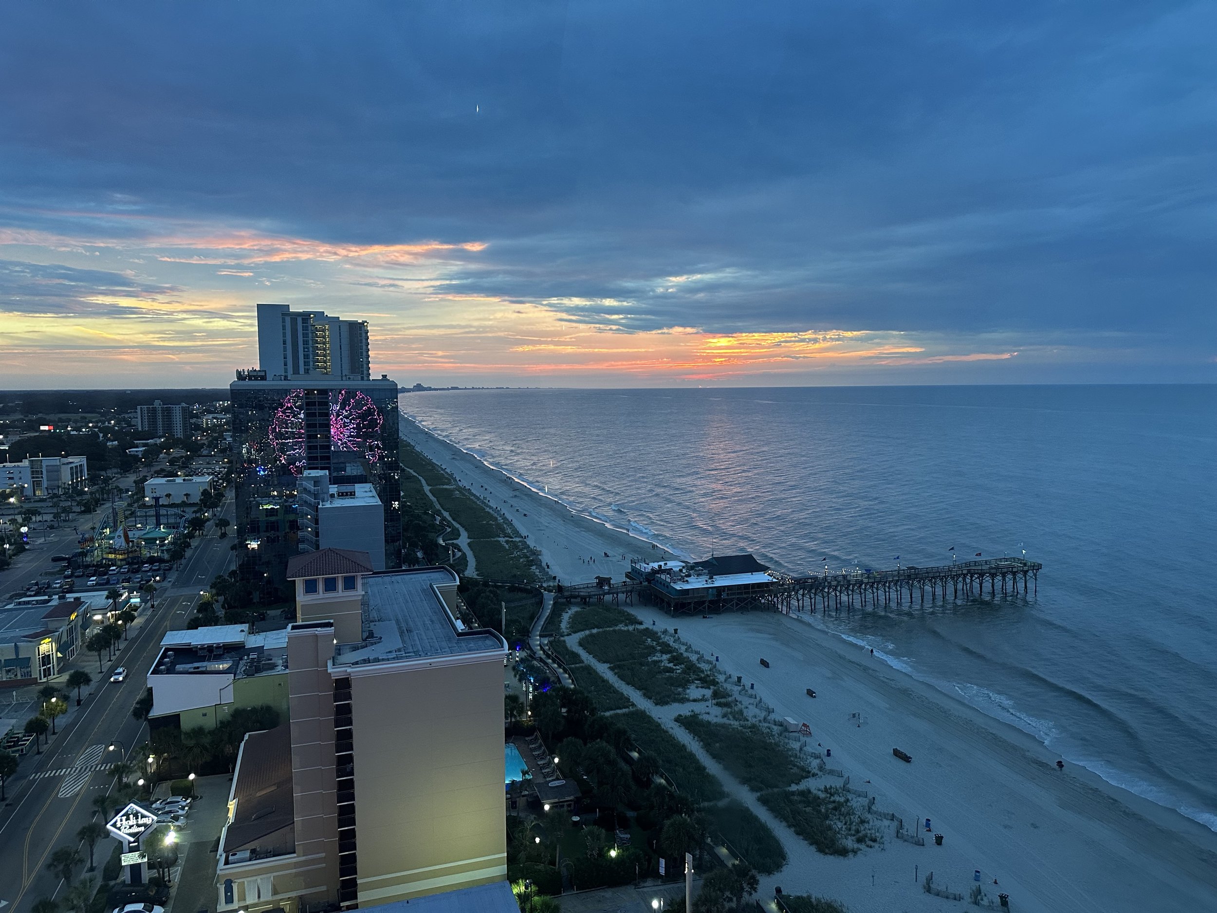 Coastal cityscape at sunrise with buildings, illuminated Ferris wheel, beachfront, and pier extending into the ocean.