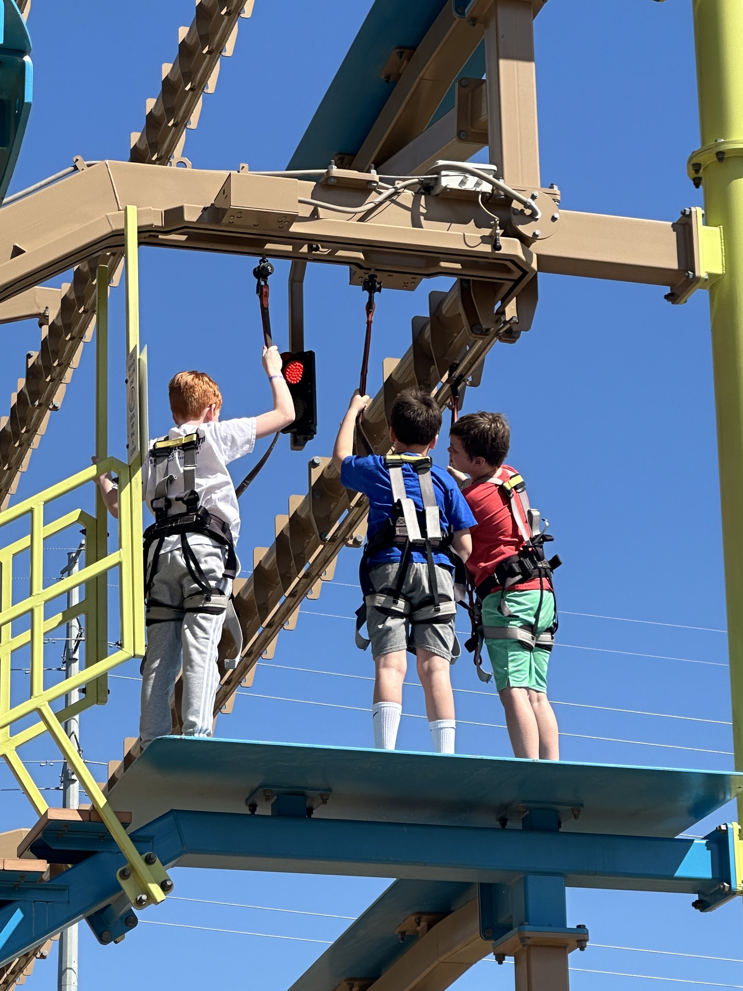 Three children wearing harnesses on a ropes course, standing on a platform, interacting with a red light signal.