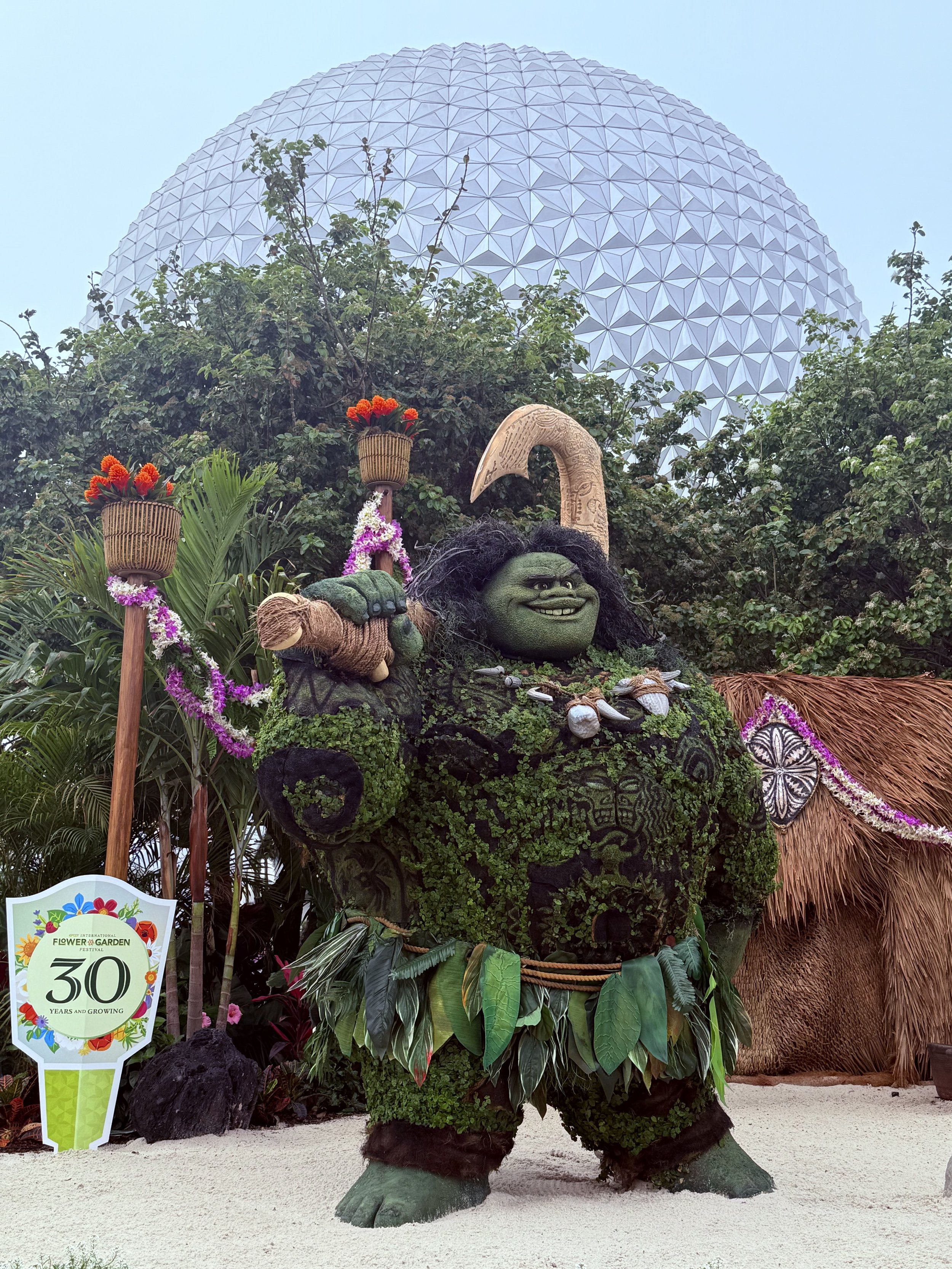 Topiary of a character with greenery and leaves, holding a staff and wearing a grass skirt. A large geodesic sphere is visible in the background, surrounded by trees. A sign reads 'Flower & Garden 30 Years and Growing.'