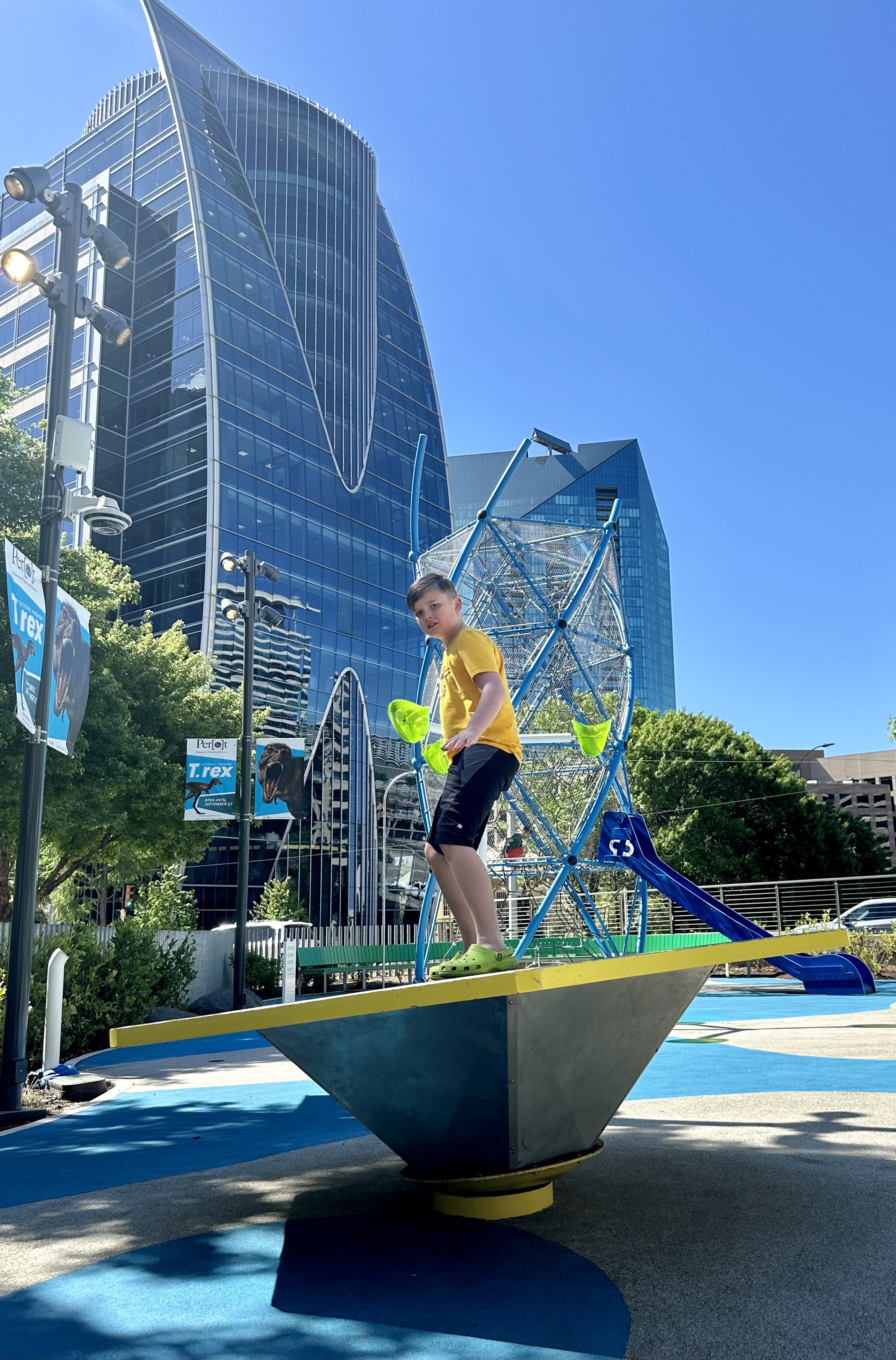 Child balancing on playground equipment in urban park with modern buildings in background.