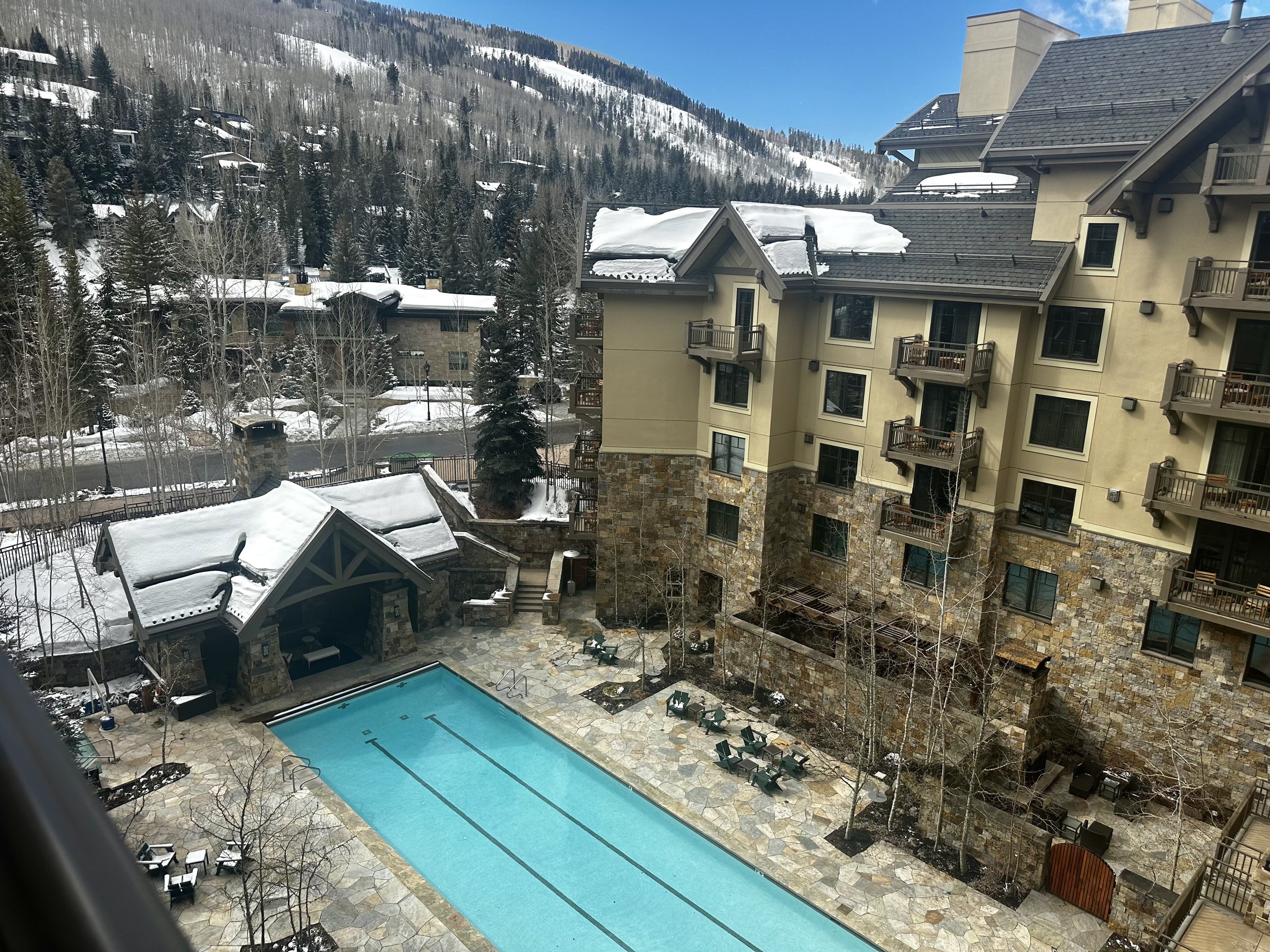 Aerial view of a resort with an outdoor swimming pool surrounded by snow-covered buildings and trees. The building has balconies and a stone facade. Snow-covered mountains and evergreen trees are visible in the background under a blue sky.
