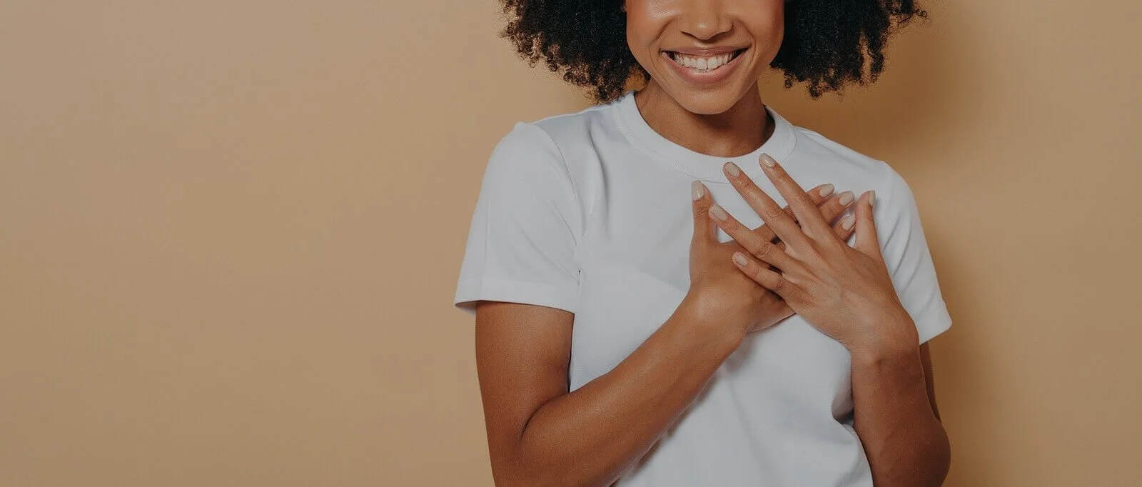 A woman smiling and placing her hands on her chest, wearing a white t-shirt against a beige background.