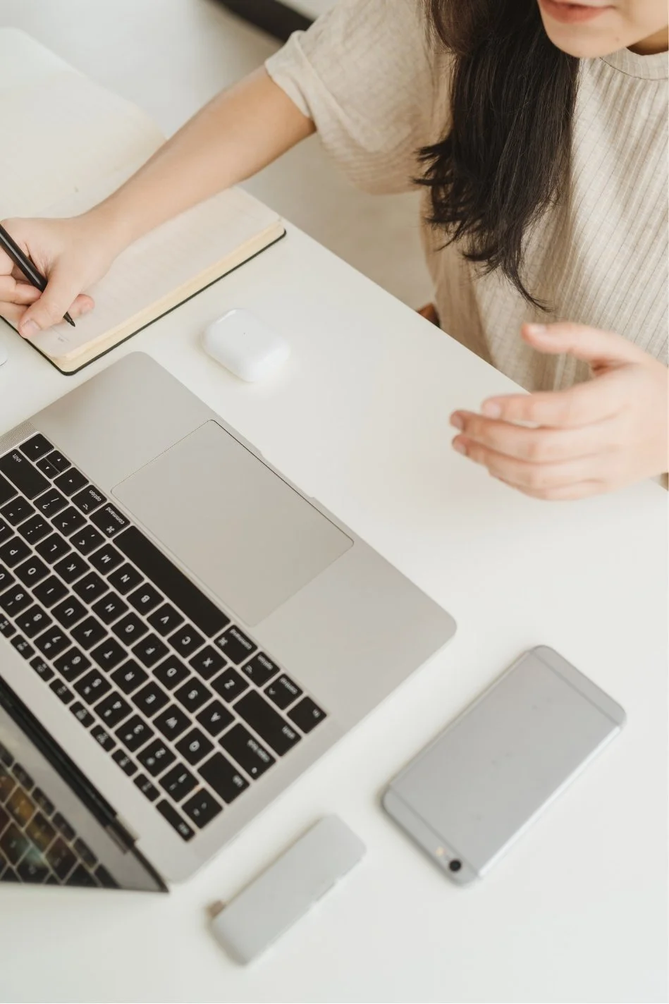 A woman with long dark hair writing in a notebook at a white desk with a laptop, smartphone, wireless earbuds, and a small white device.