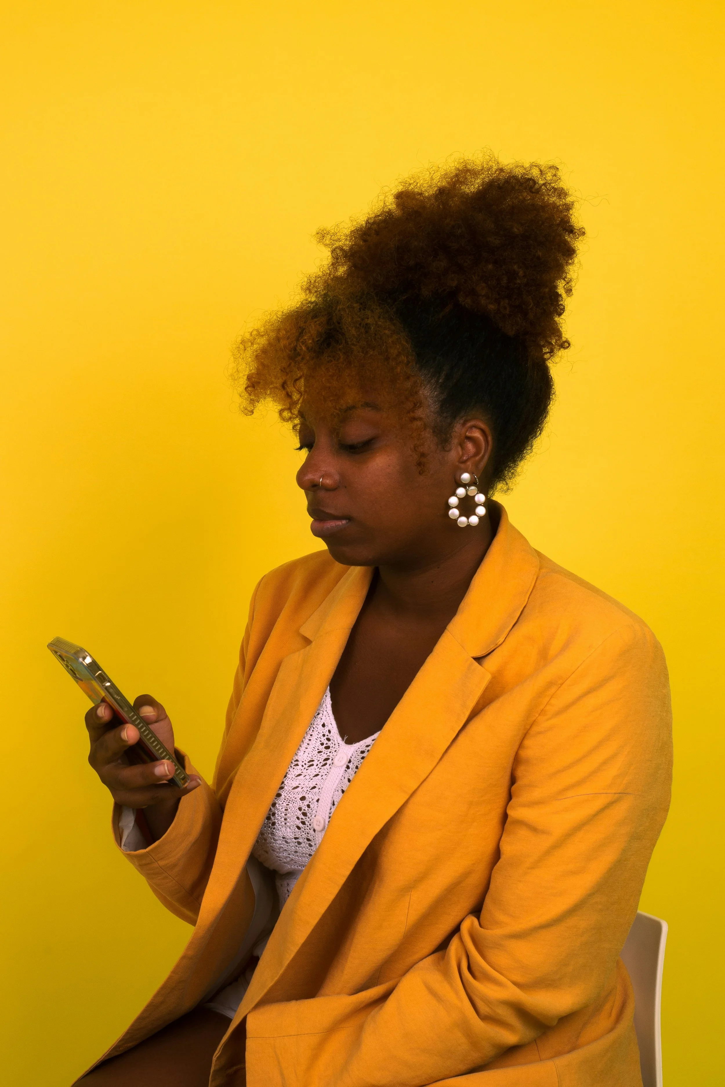 A woman with curly hair in a high puff wearing a yellow blazer and white crochet top, looking at her phone against a yellow background.