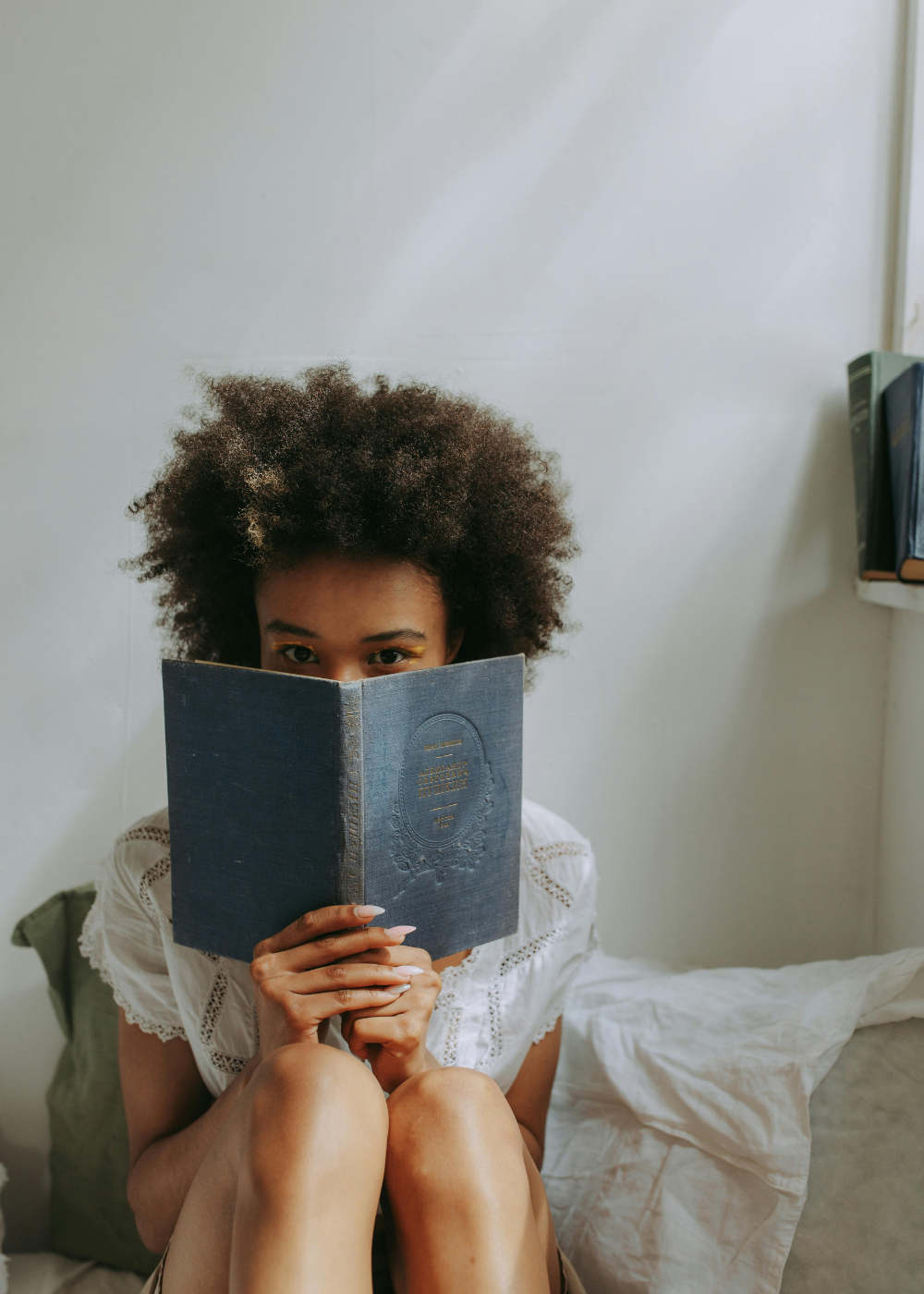 A woman with curly hair is sitting on a bed, holding a book in front of her face, with only her eyes visible.