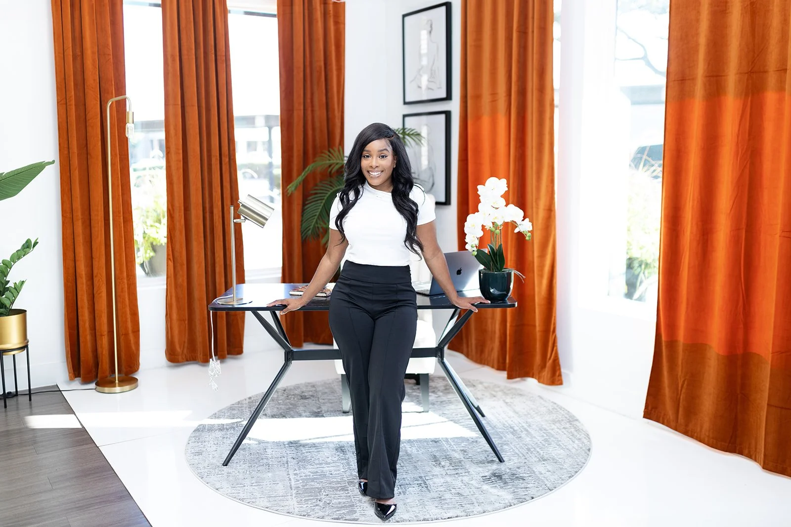 A woman in business attire posing in a modern, well-lit home office with orange curtains, a glass desk, potted plants, and wall art.