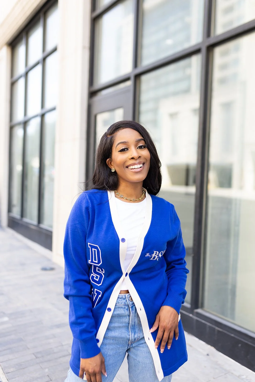 A young woman smiling outdoors near a building with large windows. She has dark hair, wears a blue baseball jacket over a white shirt, light blue jeans, and gold jewelry.