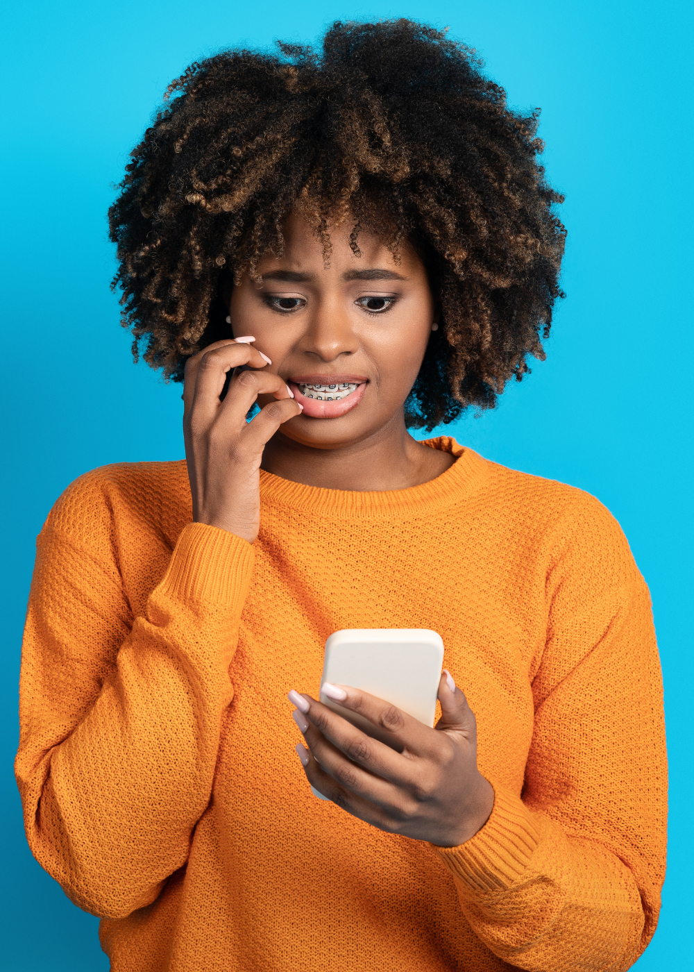 A young woman with curly hair and braces on her teeth looking at her phone with a surprised or concerned expression against a blue background. She is wearing an orange sweater.