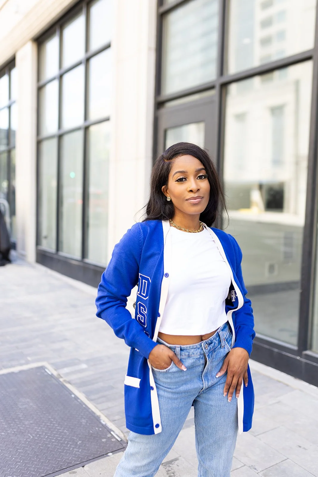 A confident woman stands outdoors in front of a modern glass building, wearing a blue letterman jacket, white crop top, and light blue jeans.