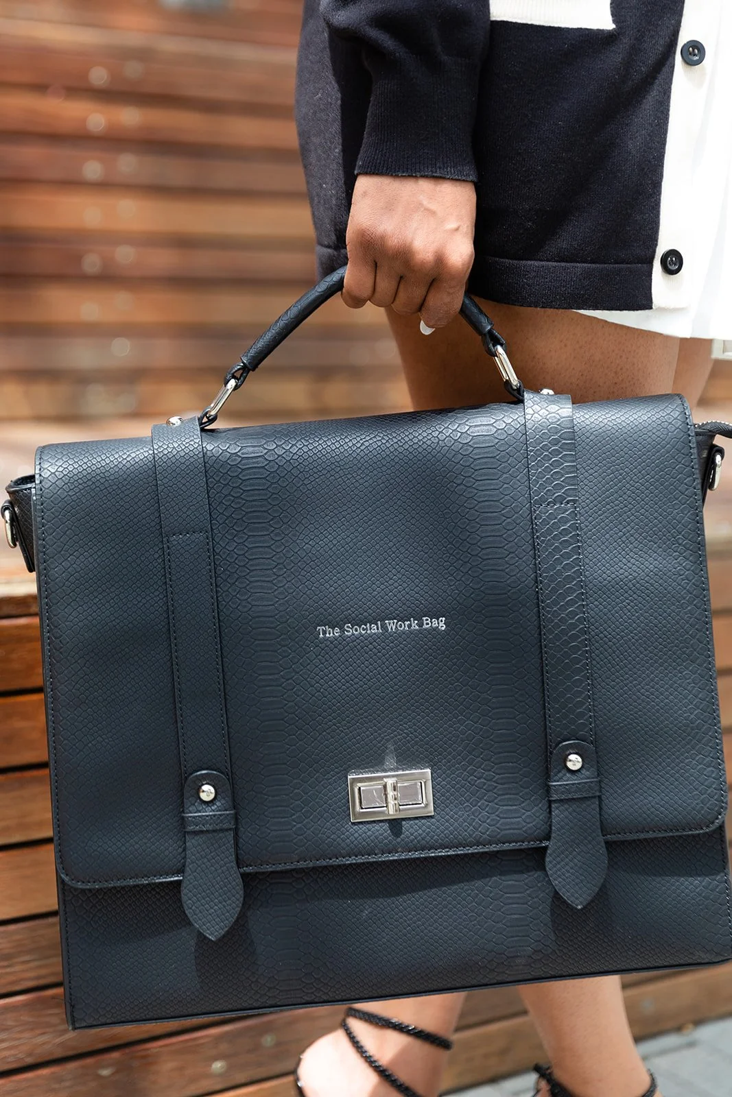 A person holding a black textured leather briefcase with a handle. The briefcase has the text 'The Social Work Bag' printed on it and features a metal clasp. The person is standing on a wooden surface.