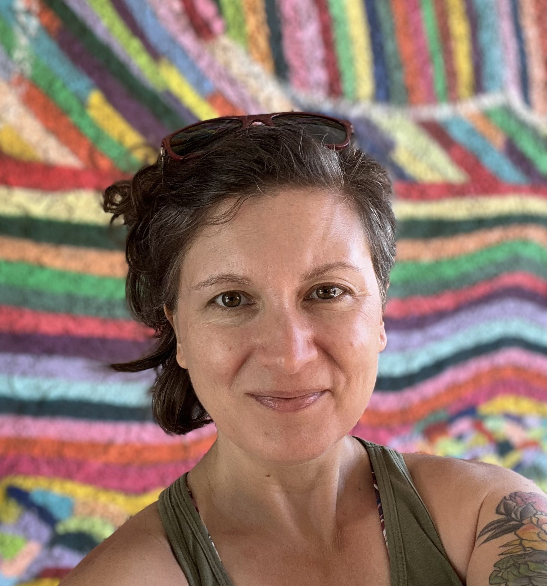 A picture of post-evangelical pastor Brenna Rubio, a smiling woman with short, dark, curly hair and glasses on her head smiling at the camera, with a dynamic rainbow of stripes in the background.