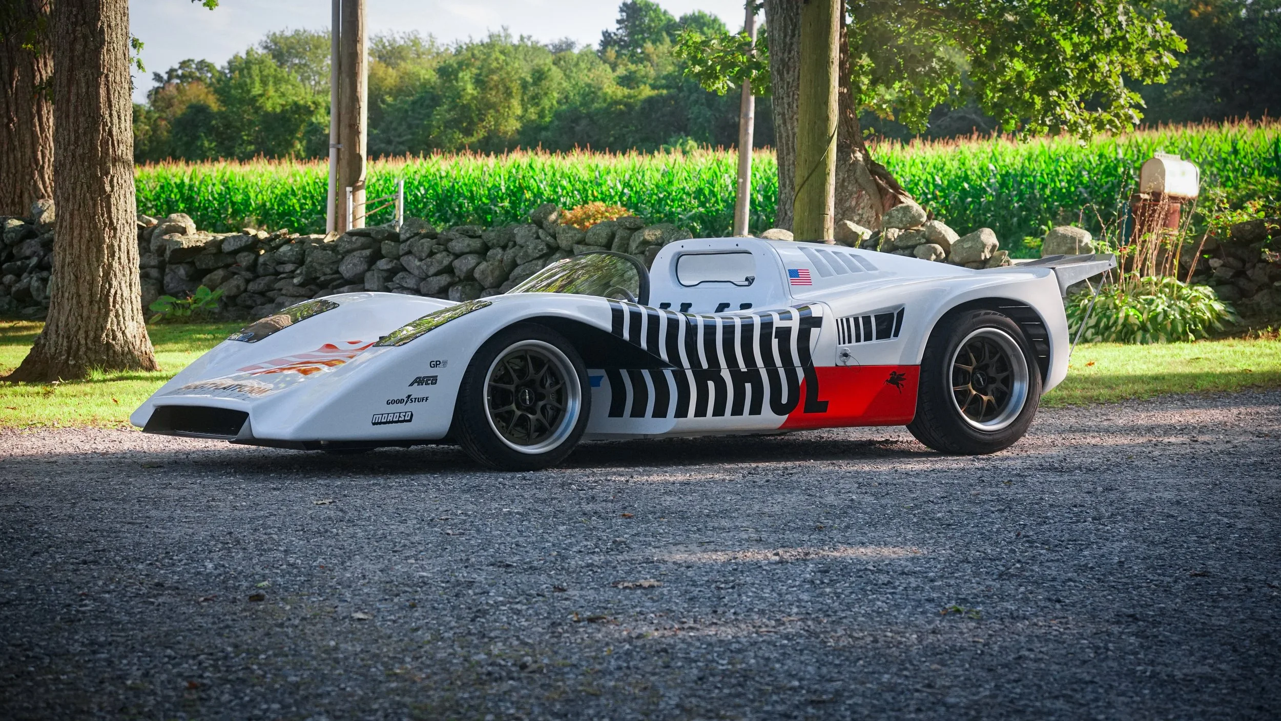 A vintage race car, white with black and orange accents, parked on a gravel surface, surrounded by trees and greenery.