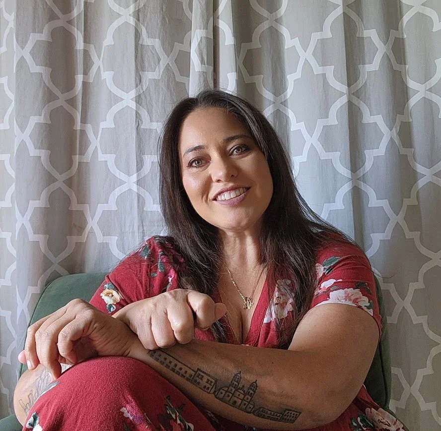 Smiling woman with dark hair, wearing a floral red dress, sitting in front of patterned curtains, showing a tattoo of a cityscape on her forearm.