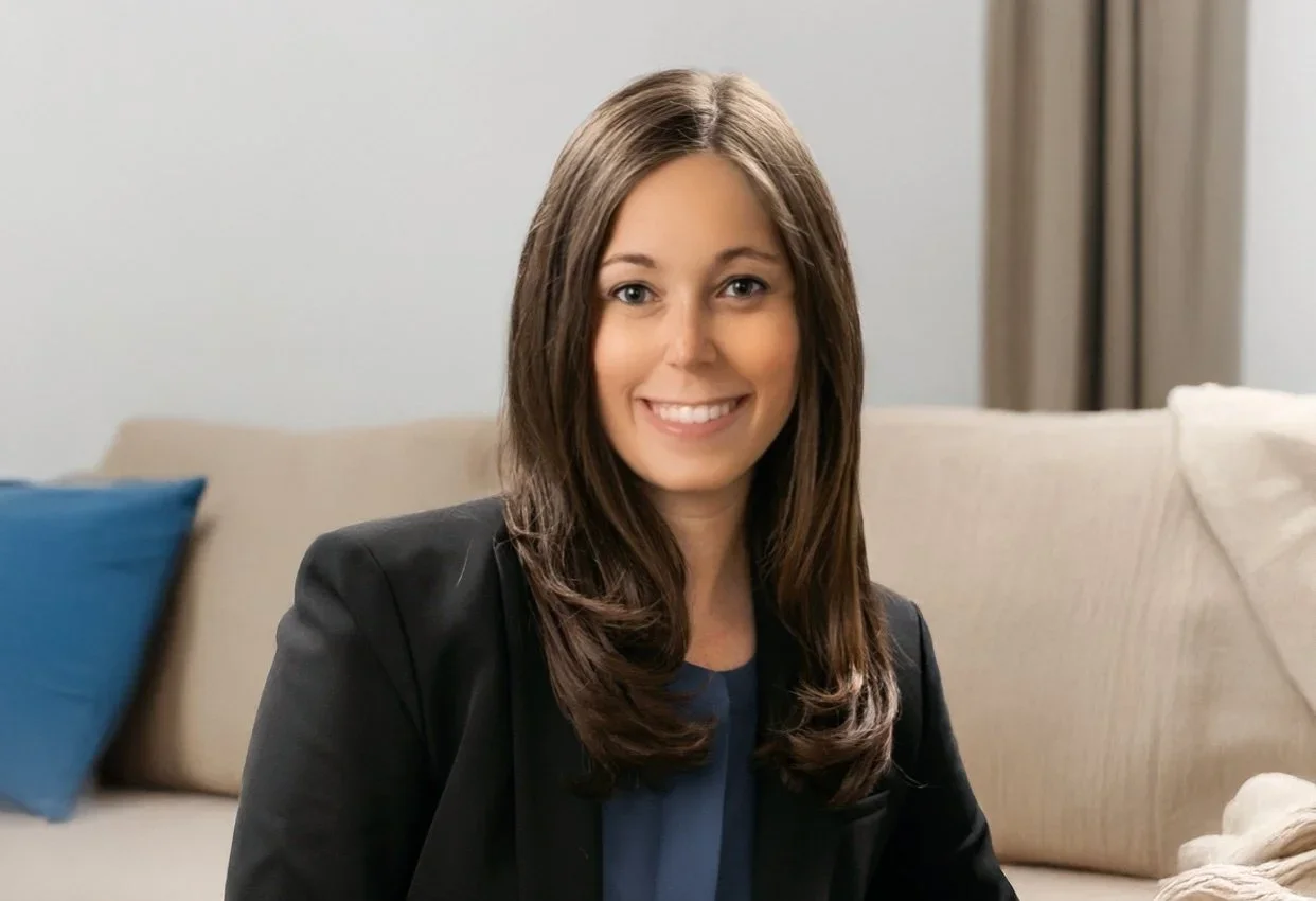 A woman with long brown hair, smiling, wearing a black blazer, sitting on a beige sofa with blue and white pillows.