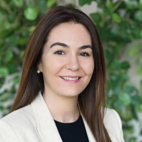 A woman with long brown hair smiling, wearing a cream blazer and pearl earrings, standing outdoors with green leafy plants in the background.