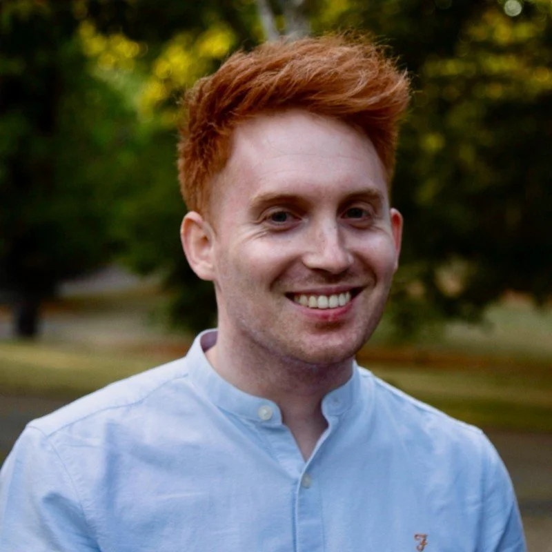 A young man with red hair and fair skin, smiling while wearing a light blue collared shirt, outdoors with trees and blurred background.