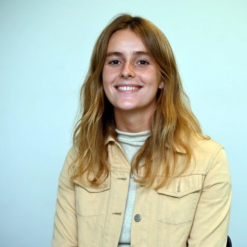 A young woman with long red hair, freckles, and a broad smile, wearing a beige jacket over a light-colored turtleneck, standing against a light-colored background.
