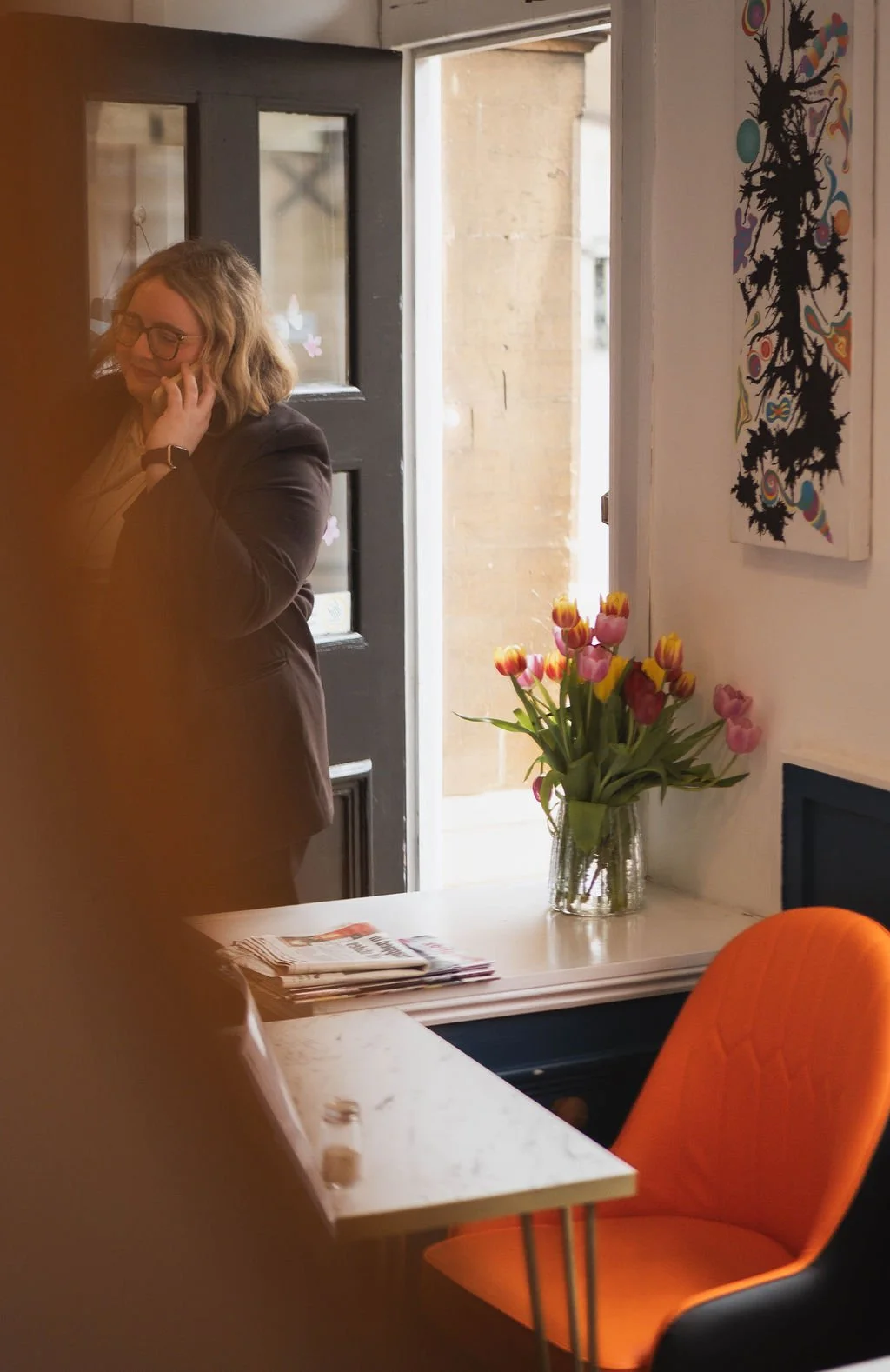 Inside a cozy cafe, a woman with glasses and shoulder-length hair is talking on her phone near the entrance, which is slightly open, revealing a stone building outside. A vase of pink and yellow tulips sits on a white surface, with a colorful abstract artwork hanging on the wall. There is an orange chair and some magazines on the table.