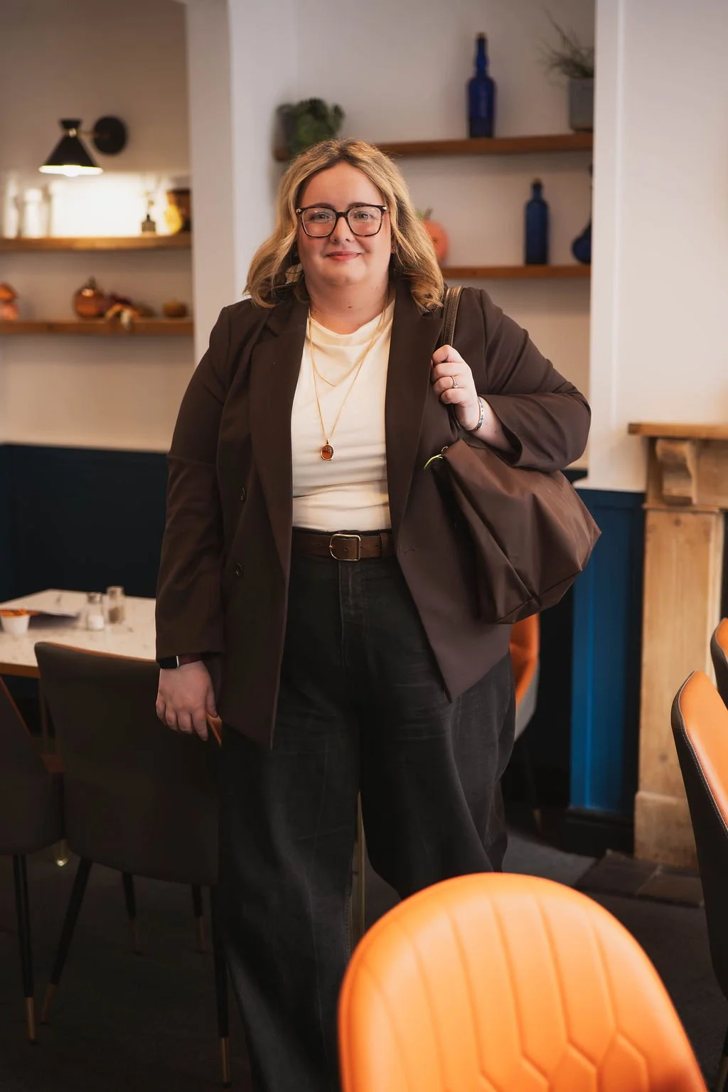 A woman with glasses and shoulder-length blonde hair, wearing a dark blazer, white top, and black jeans, carries a brown bag over her shoulder and stands in a restaurant or cafe.