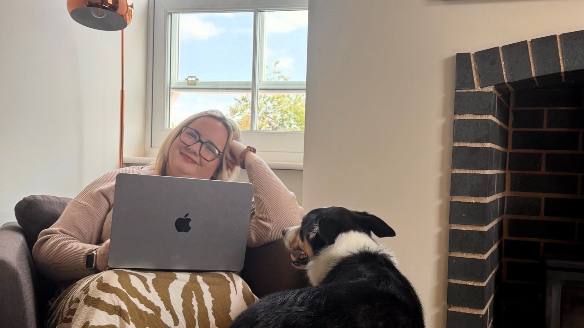 A woman with glasses and a pink sweater is lying on a sofa with her head resting on her hand, smiling at the camera. She has a laptop in front of her and a black and white dog sitting nearby, looking up at her. The room has a window with a view of a blue sky and a brick fireplace.