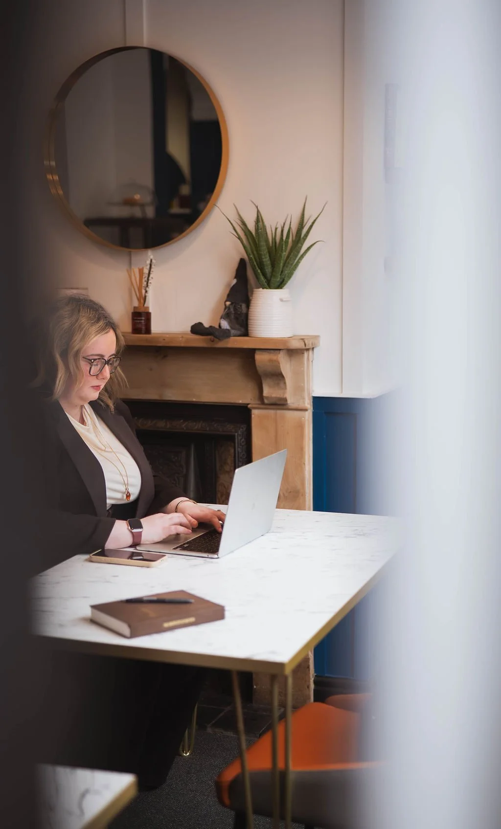 A woman with blonde hair and glasses working on a laptop at a white marble table, with a notebook, phone, and pen nearby, in a cozy room with a mirror, plants, and wooden decor.