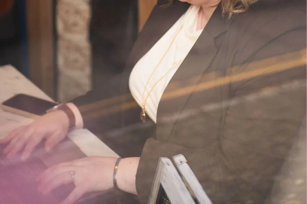 A woman sitting at a table, writing in a notebook, with a smartphone nearby, seen through a glass window.