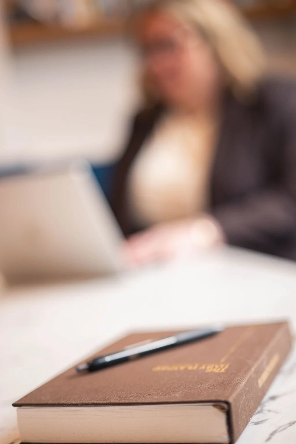 A closed brown hardcover book with a pen resting on top, placed on a white marble surface in front of a blurred woman sitting at a desk.