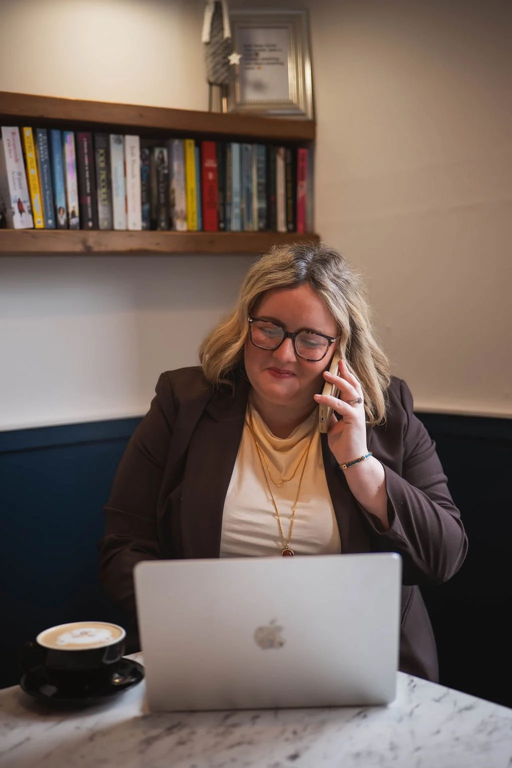 A woman with shoulder-length blonde hair and glasses sitting at a table in a cafe, talking on her smartphone, with a black cup of coffee and an open MacBook in front of her, and a wooden bookshelf and framed certificate on the wall behind her.