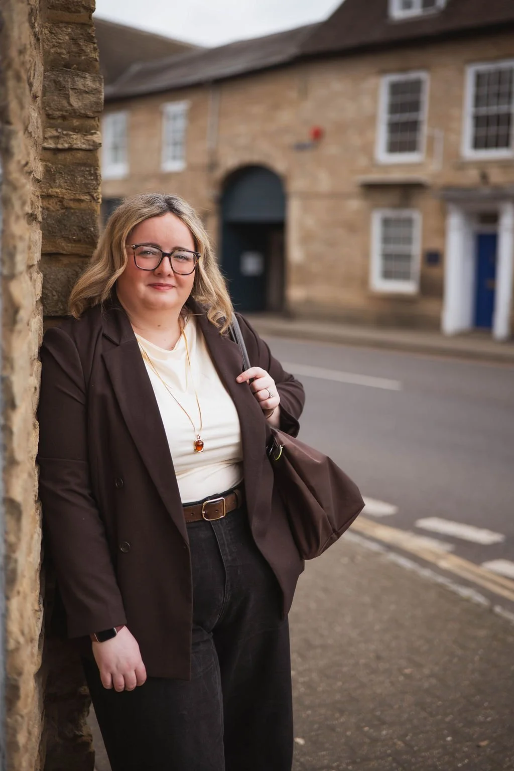 A woman with blonde hair, glasses, and a brown blazer standing on a street, leaning against a stone building with a slight smile, carrying a brown bag over her shoulder.