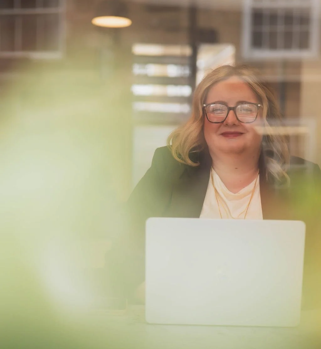 A woman with glasses and blonde hair sitting at a desk using a laptop, seen through a glass window with reflections and a blurred green foreground.