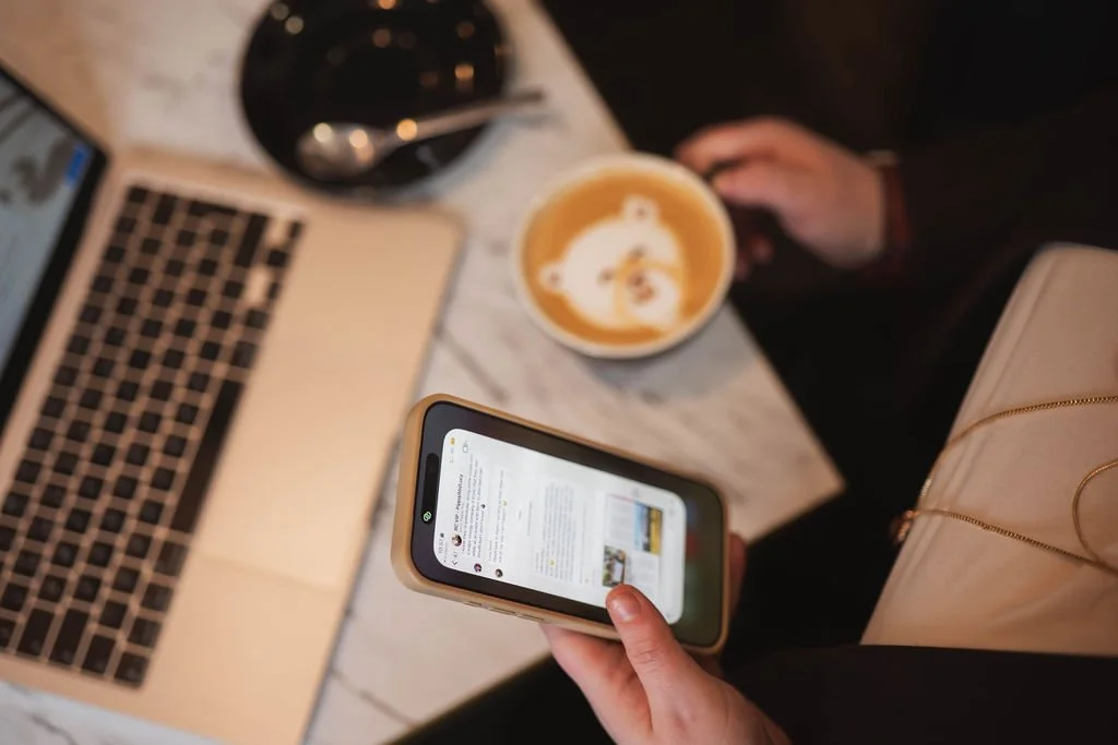Top-down view of a person holding a smartphone with a laptop, coffee, and a glass on a white marble table.