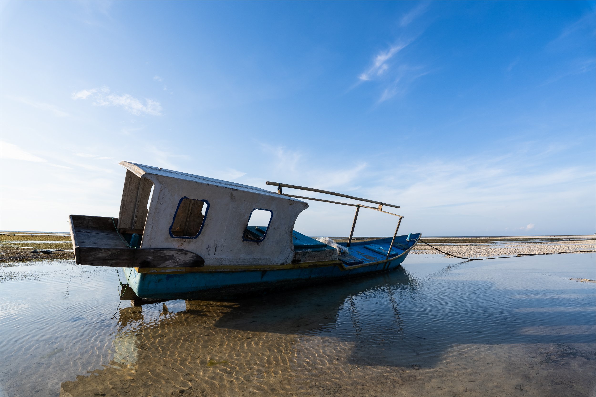 An abandoned boat resting on shallow water with a sandy shore in the background under a blue sky.
