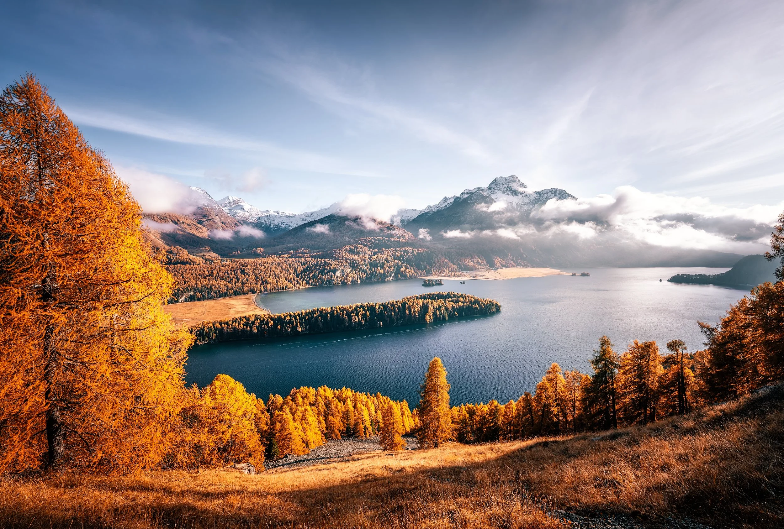 Scenic landscape of a large lake surrounded by orange and yellow autumn trees with snow-capped mountains and a partly cloudy sky in the background.