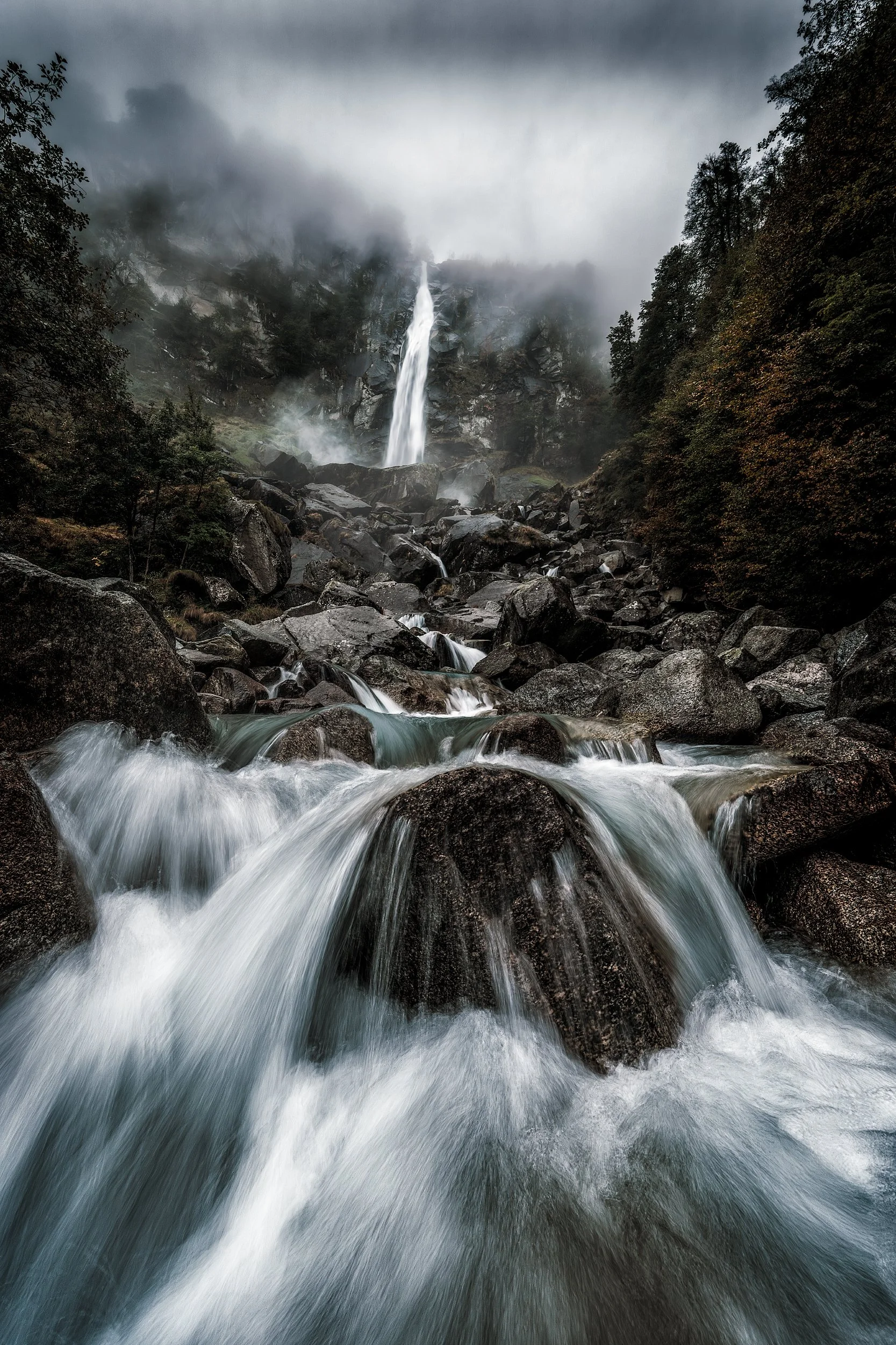 Misty mountain waterfall cascading over rocks into a rushing stream, surrounded by dark green trees and clouds overhead.