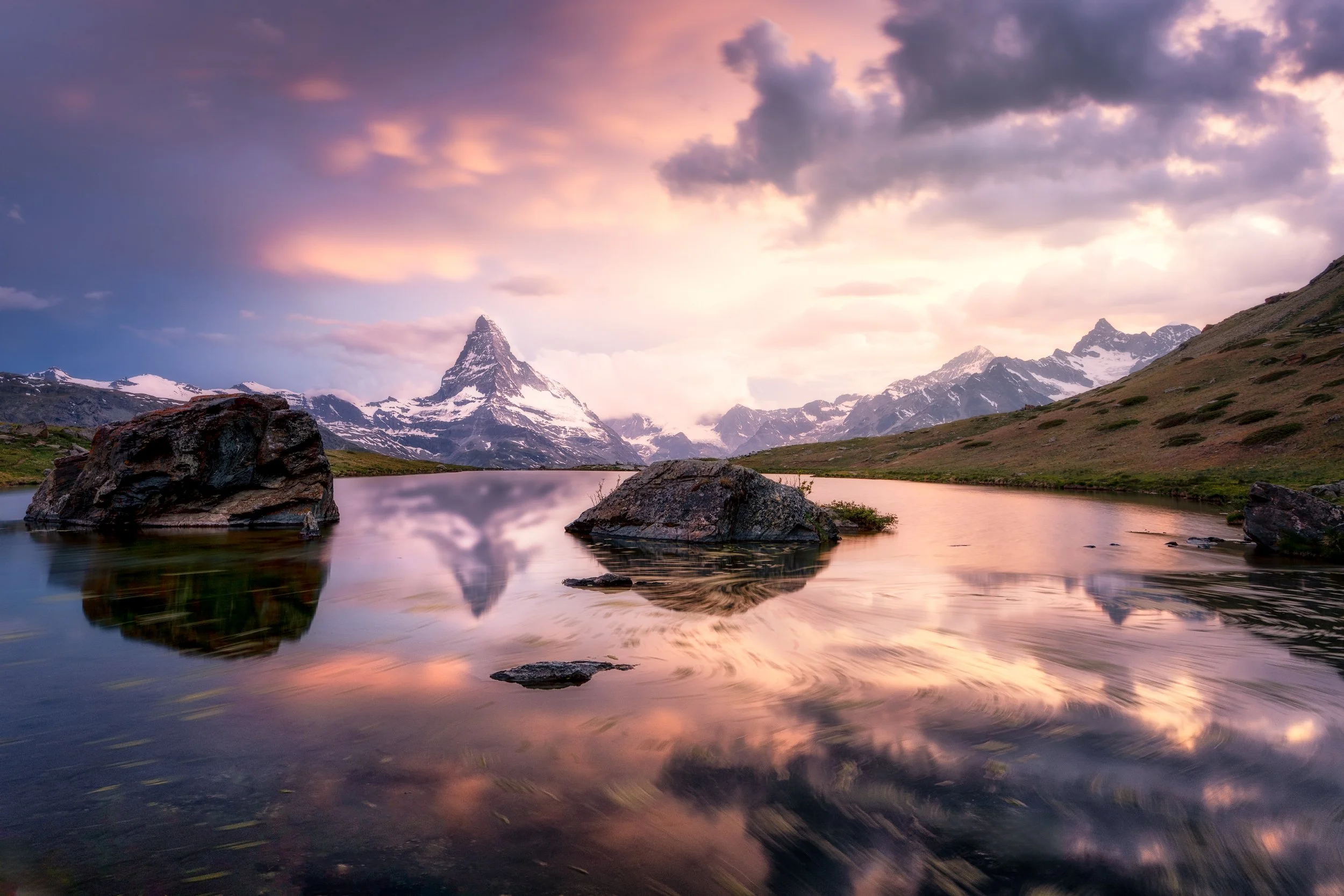 Scenic landscape of a mountain lake with snow-capped peaks in the distance, large rocks in the water, and a colorful, cloudy sky reflecting on the calm surface of the lake.