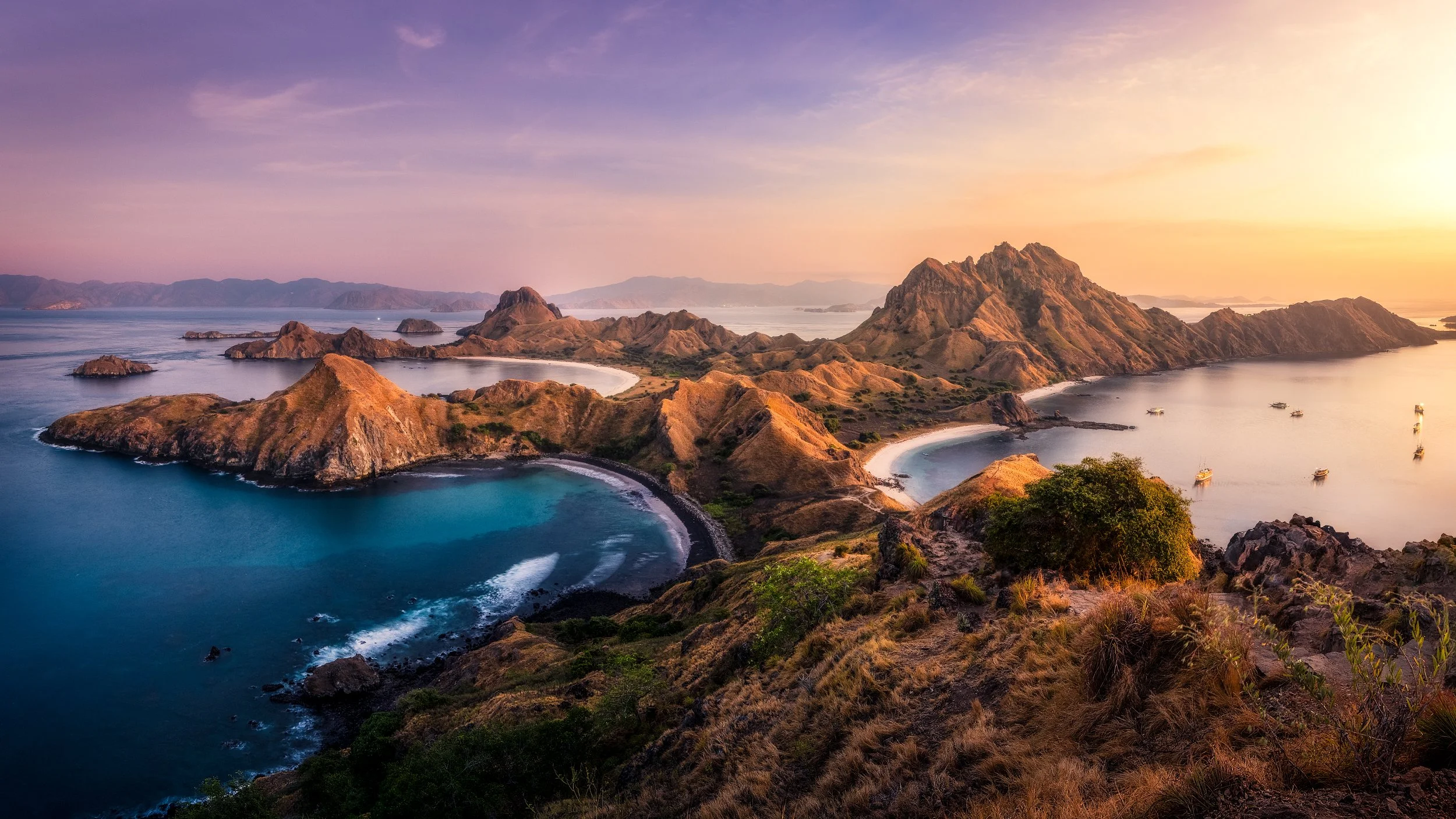 A panoramic view of a coastal landscape at sunset with rugged hills, small islands, a bay with a beach, and boats anchored in the water.