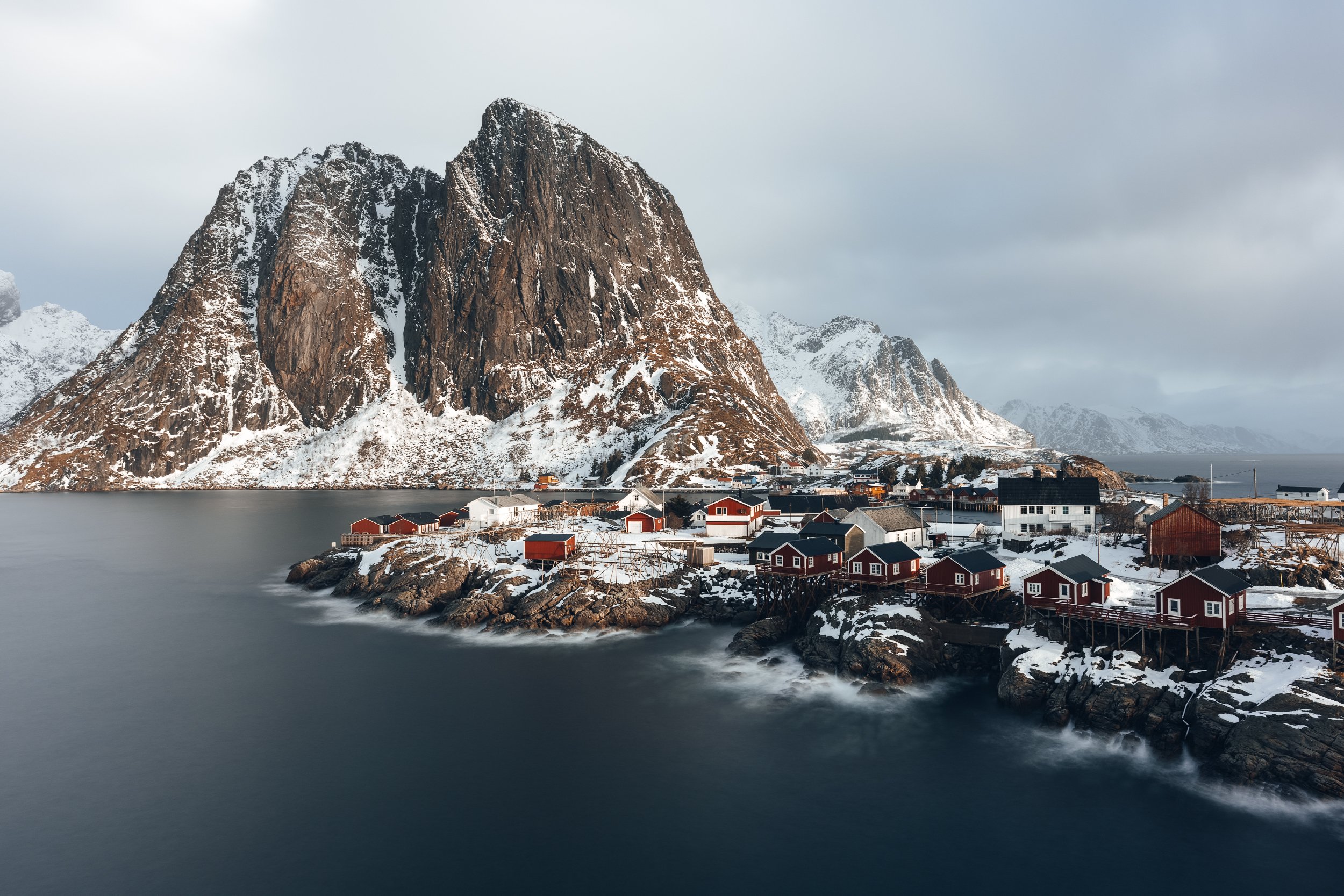 Scenic view of a Norwegian fjord with a large mountain, snow-covered houses on rocky coastline, and water in the foreground during overcast weather.
