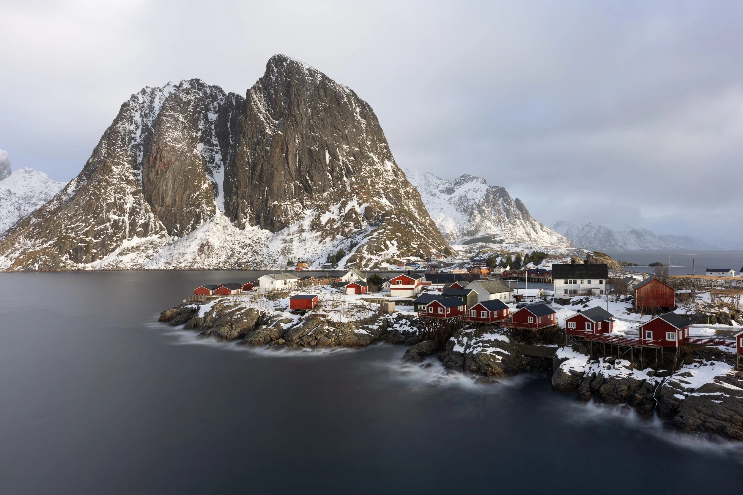 A coastal village with snow-covered ground and red cabin houses along the shoreline, with towering snow-dusted mountains in the background and calm water in the foreground.
