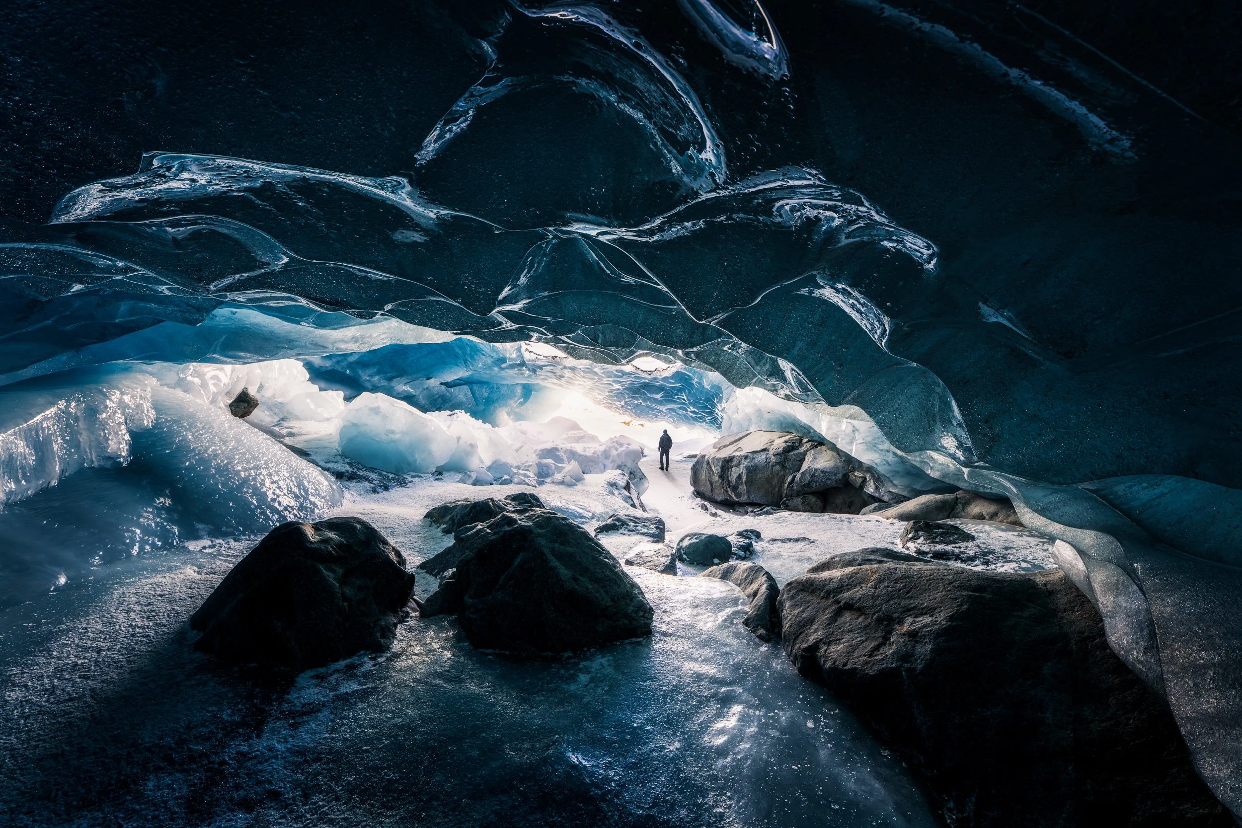 A person standing inside an ice cave with ice formations on the ceiling and ice chunks on the floor, surrounded by dark rock and snow.