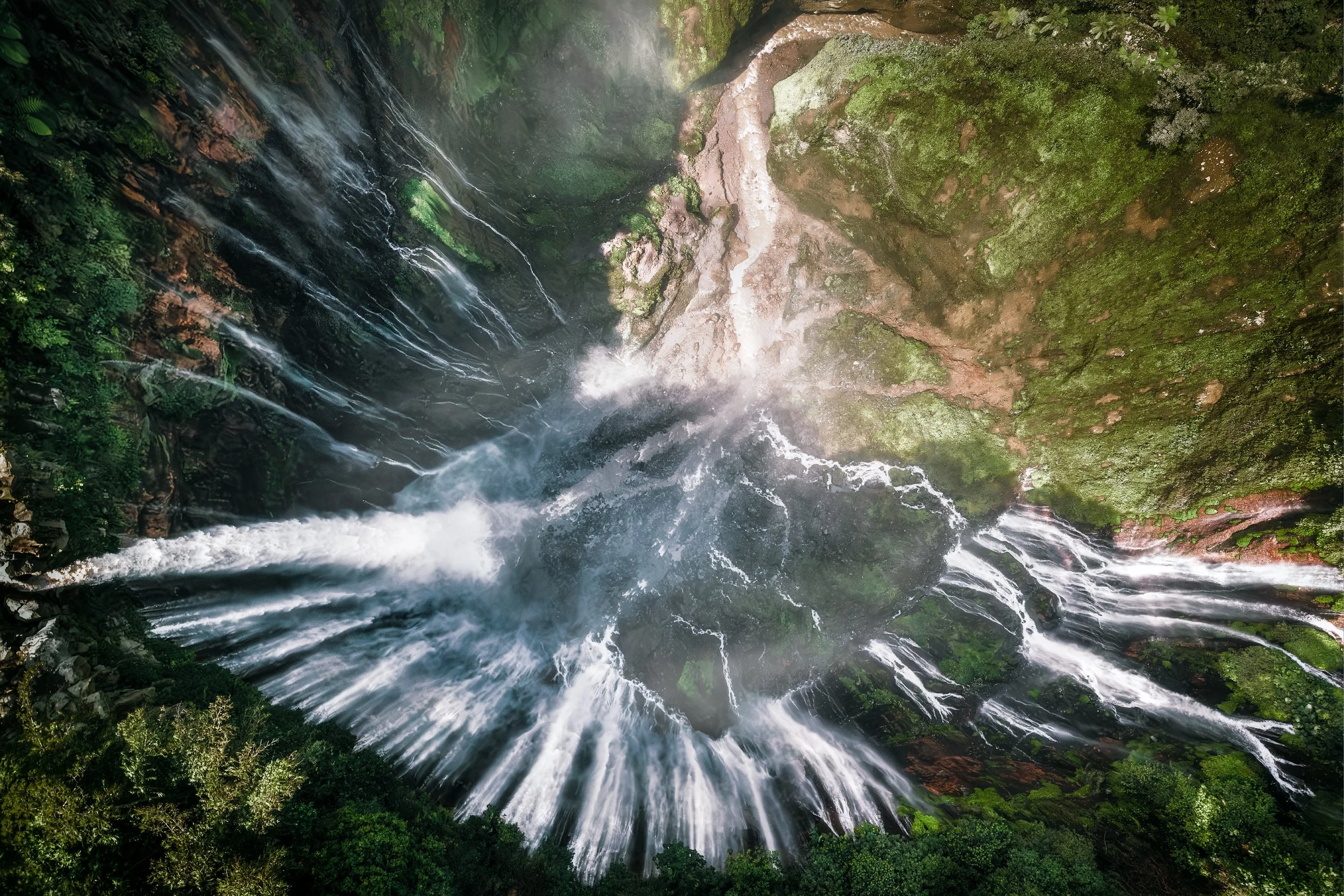 Aerial view of a waterfall cascading down mossy rocks surrounded by lush green forest.
