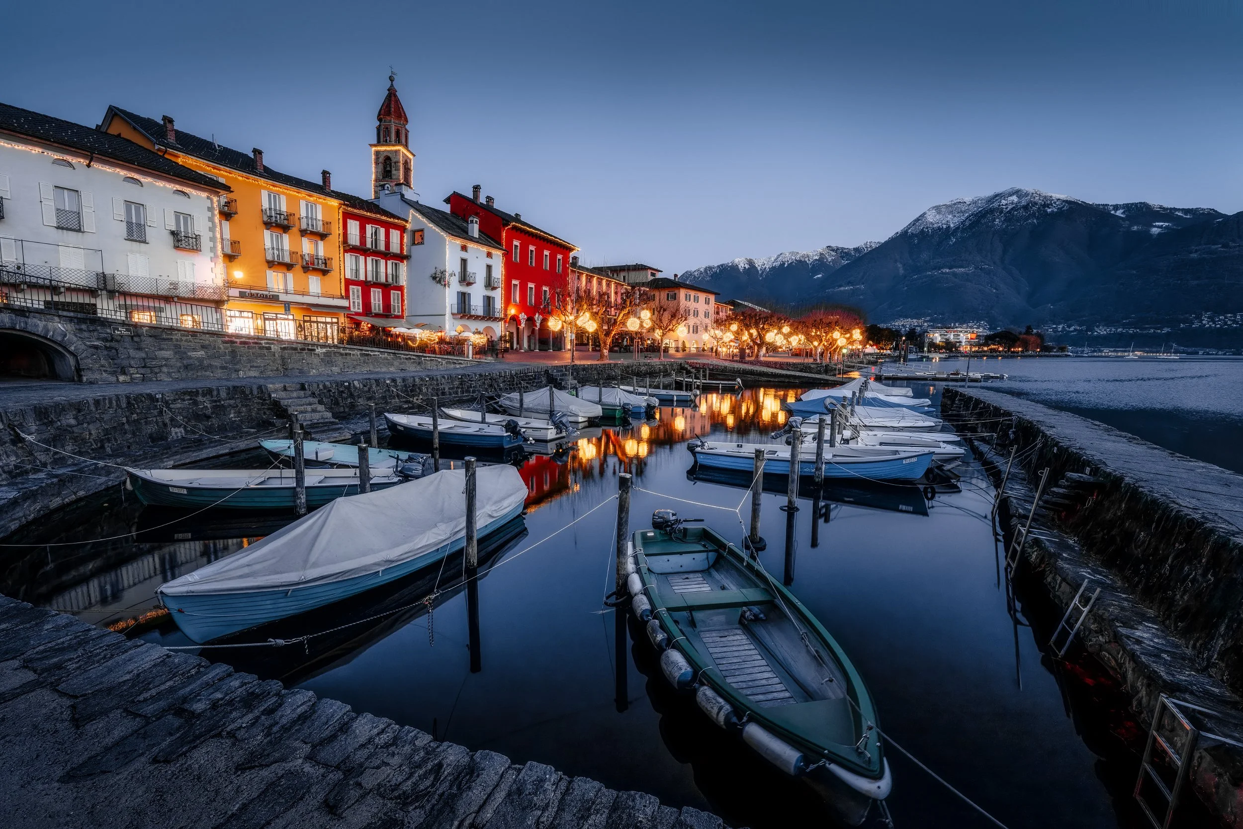 A peaceful harbor scene at dusk with small boats docked along the pier, colorful buildings with lit windows lining the waterfront, snow-capped mountains in the background, and calm reflective water.