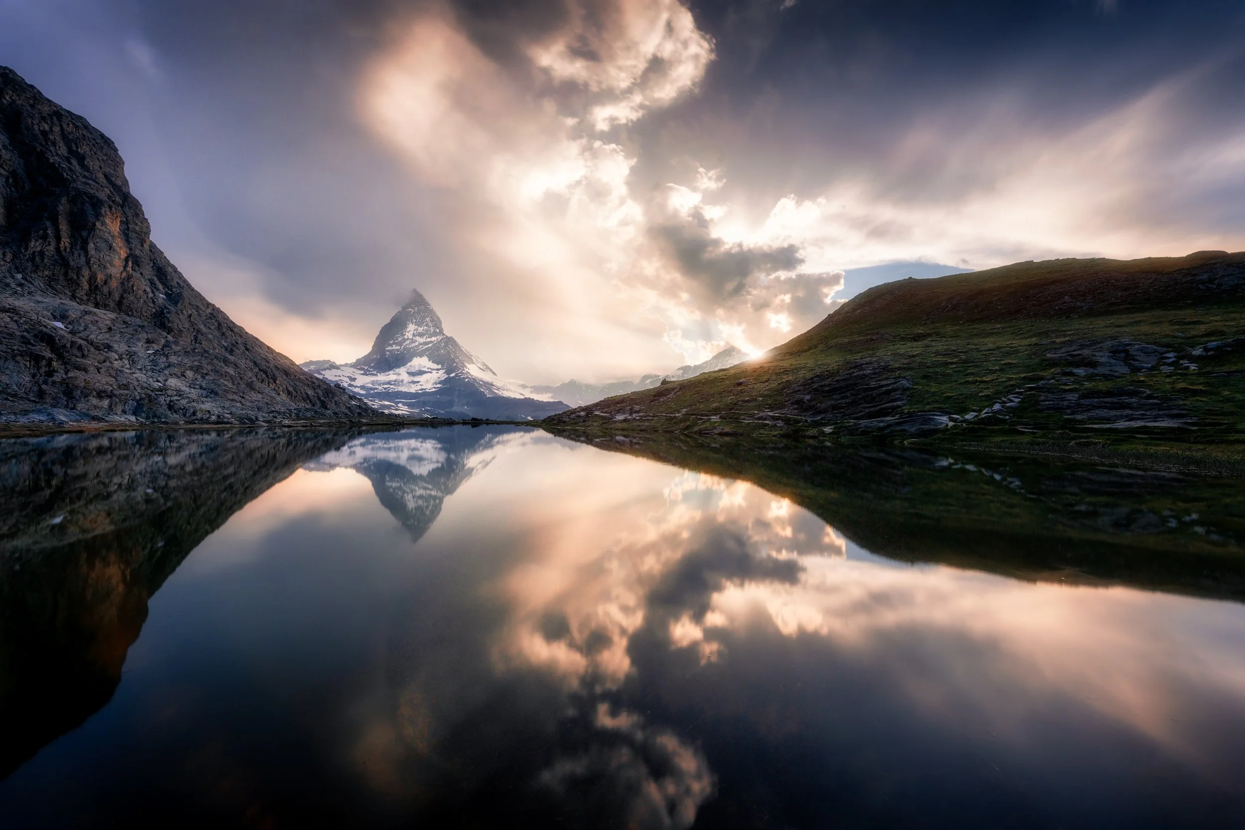 A mountain landscape with a snow-capped peak, reflecting in a calm lake. The sky has dramatic clouds with sunlight breaking through.